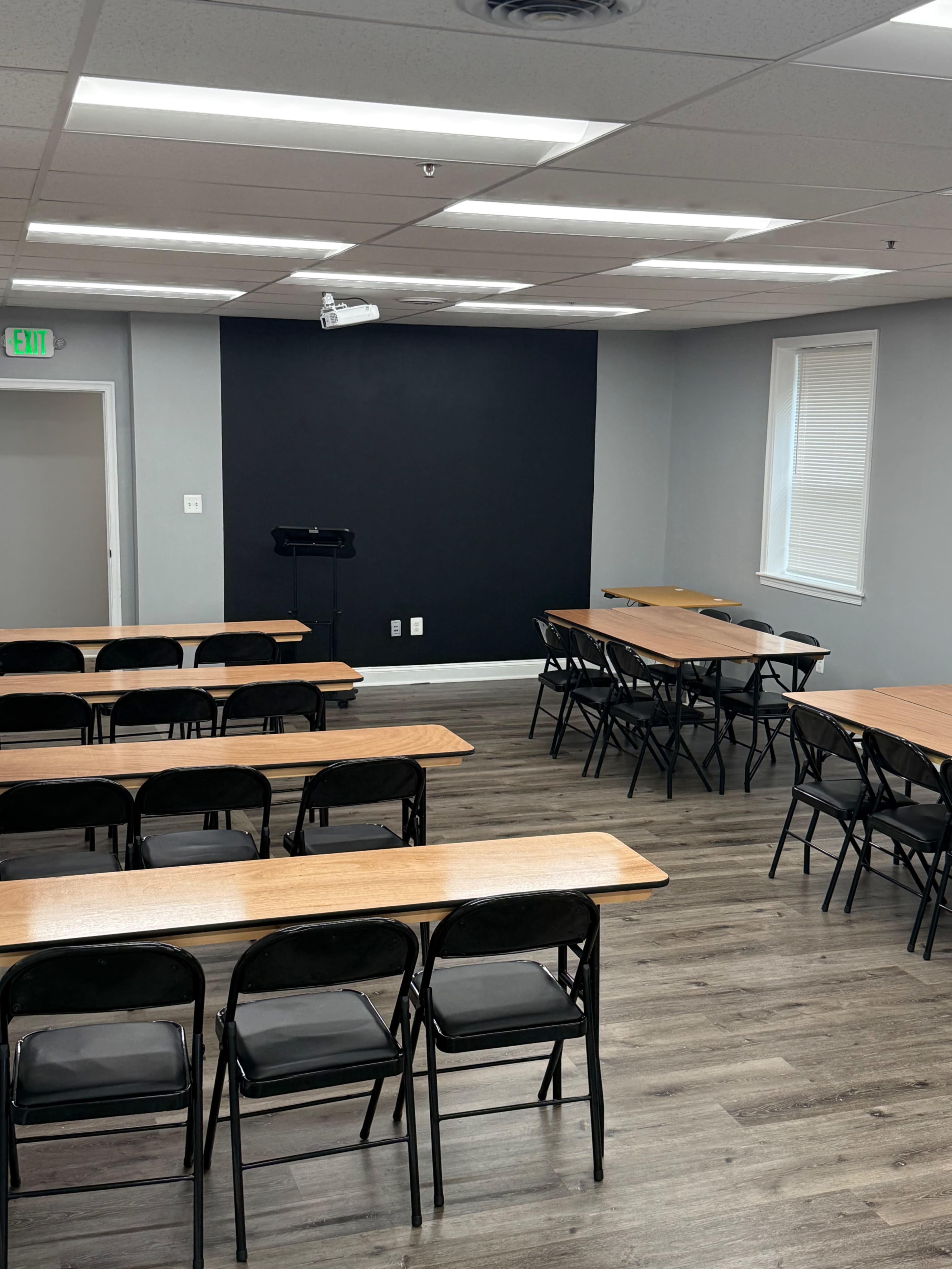 The image shows a classroom with several rows of wooden tables and black folding chairs arranged neatly, along with a projector mounted on the ceiling and a black wall at the front.