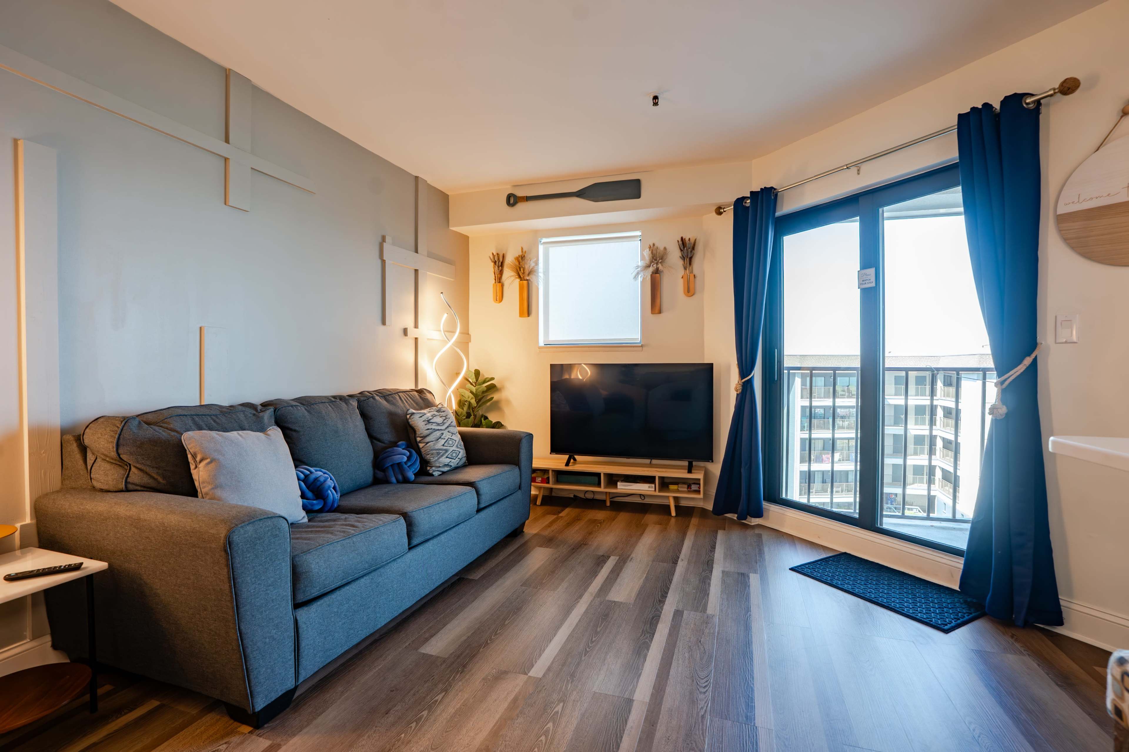 A modern living room features a gray sofa facing a television, with large windows providing natural light and access to a balcony.