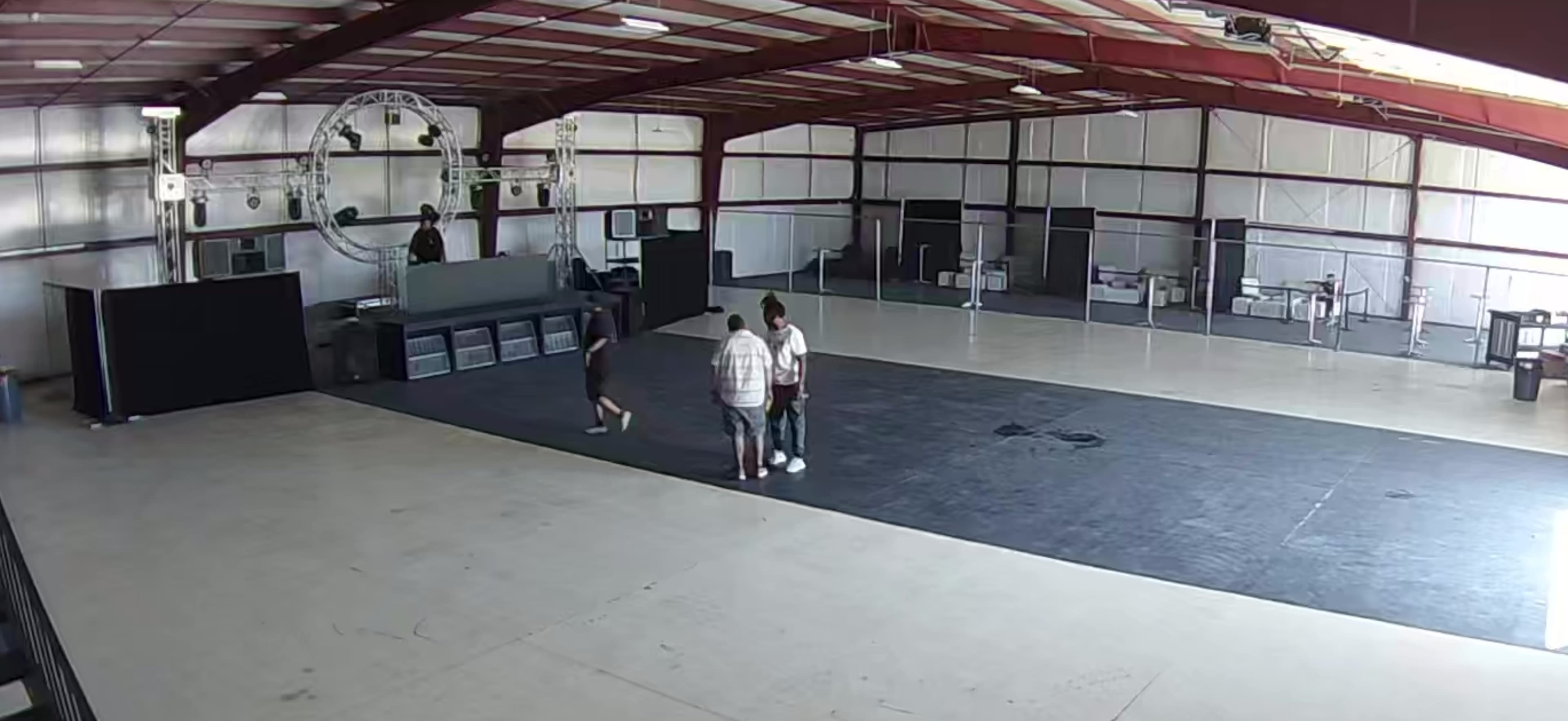 A large, empty indoor space with high ceilings features four people gathered in the center while a DJ booth is located at one end.