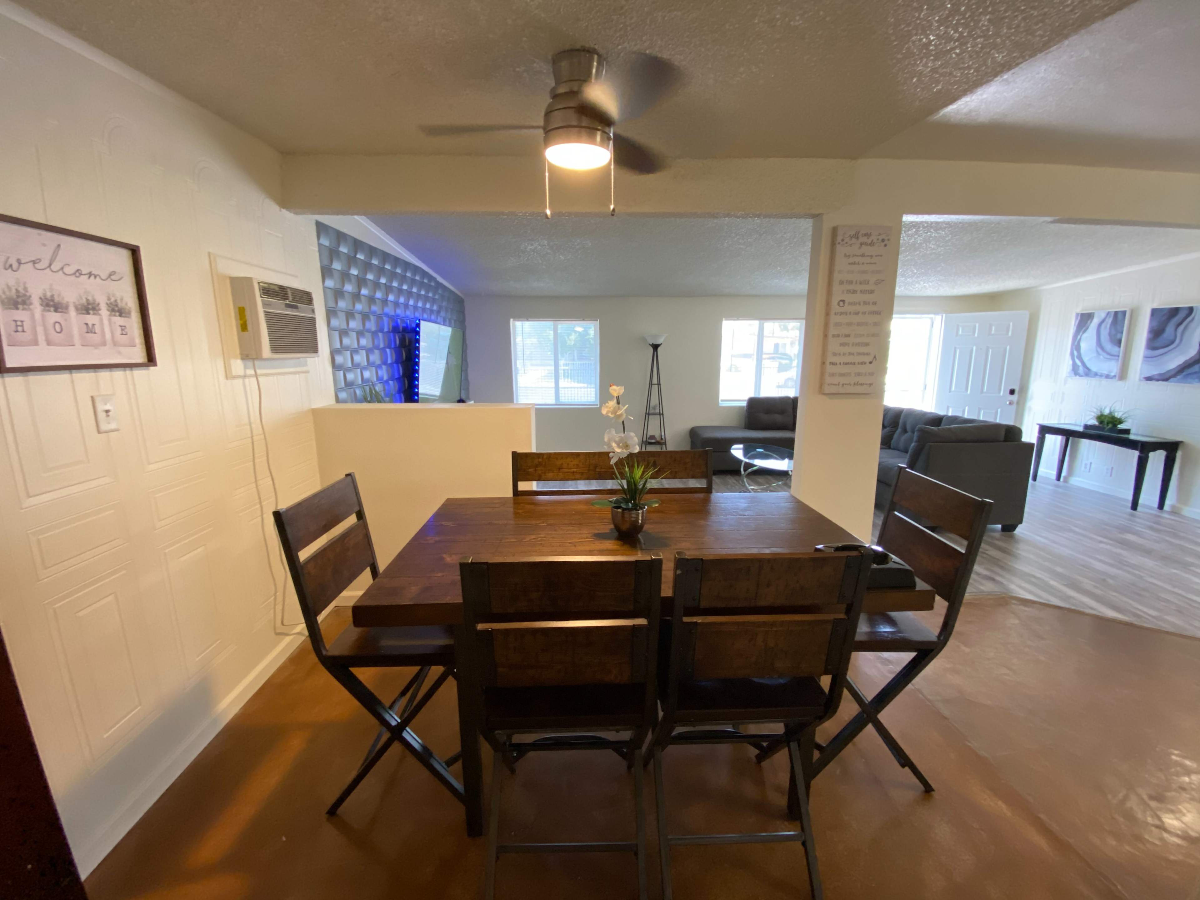 A dining area with a wooden table surrounded by folding chairs, leading into a brightly lit living space with a couch and wall-mounted TV.