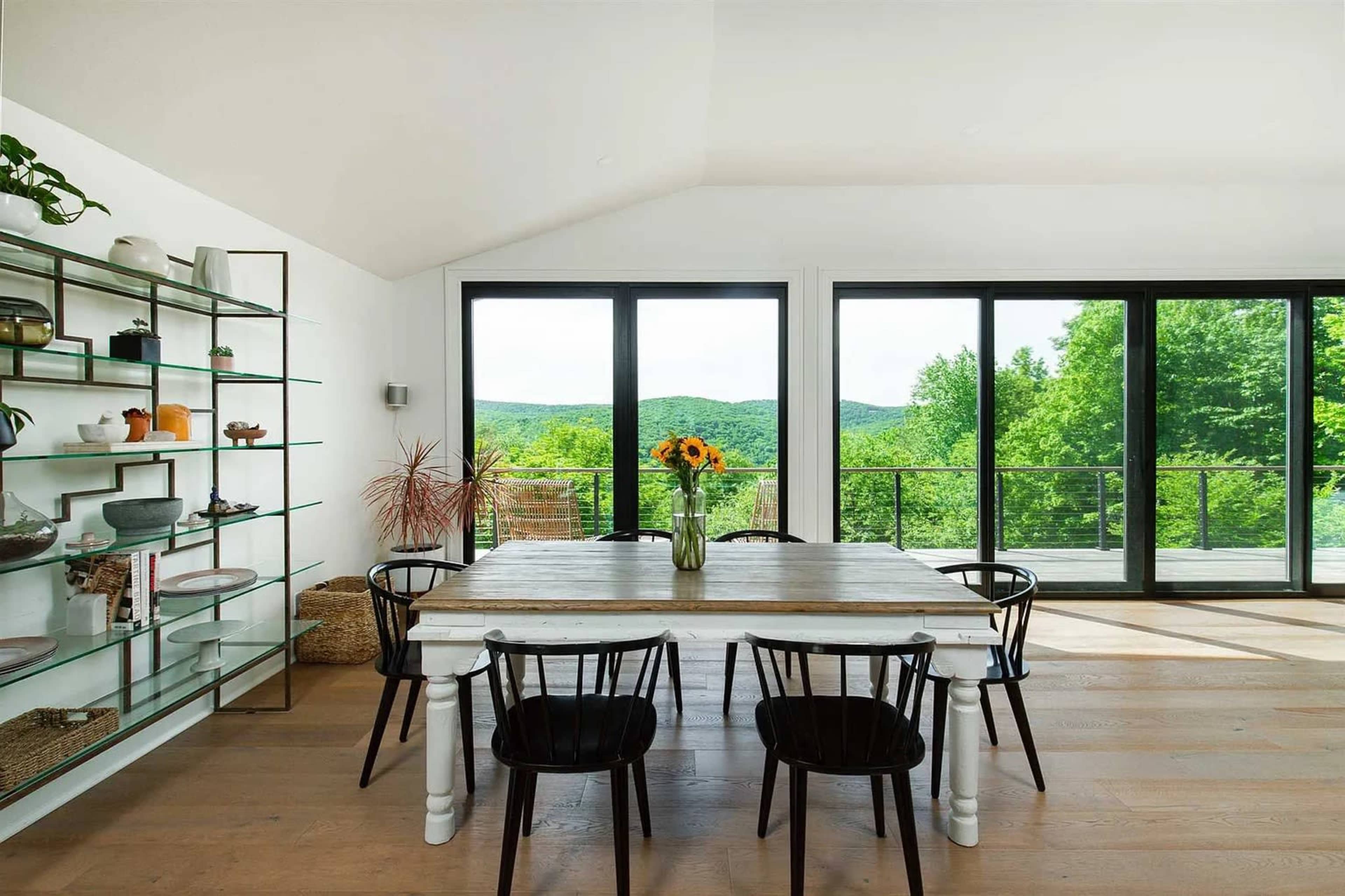 A dining area features a wooden table with black chairs, surrounded by large windows that showcase a view of green hills.