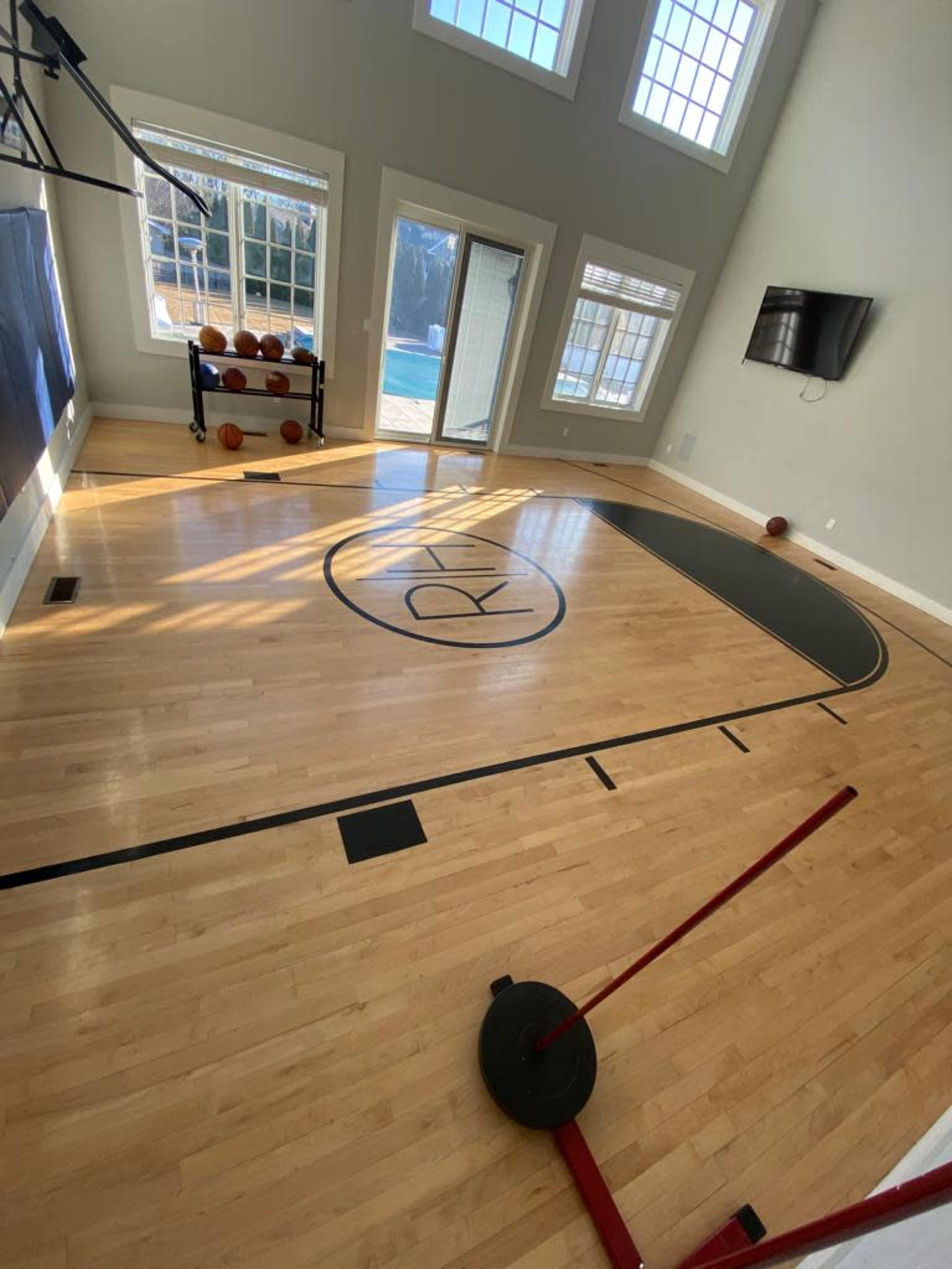 The image shows a hardwood basketball court inside a spacious room with large windows and a view of a pool outside.
