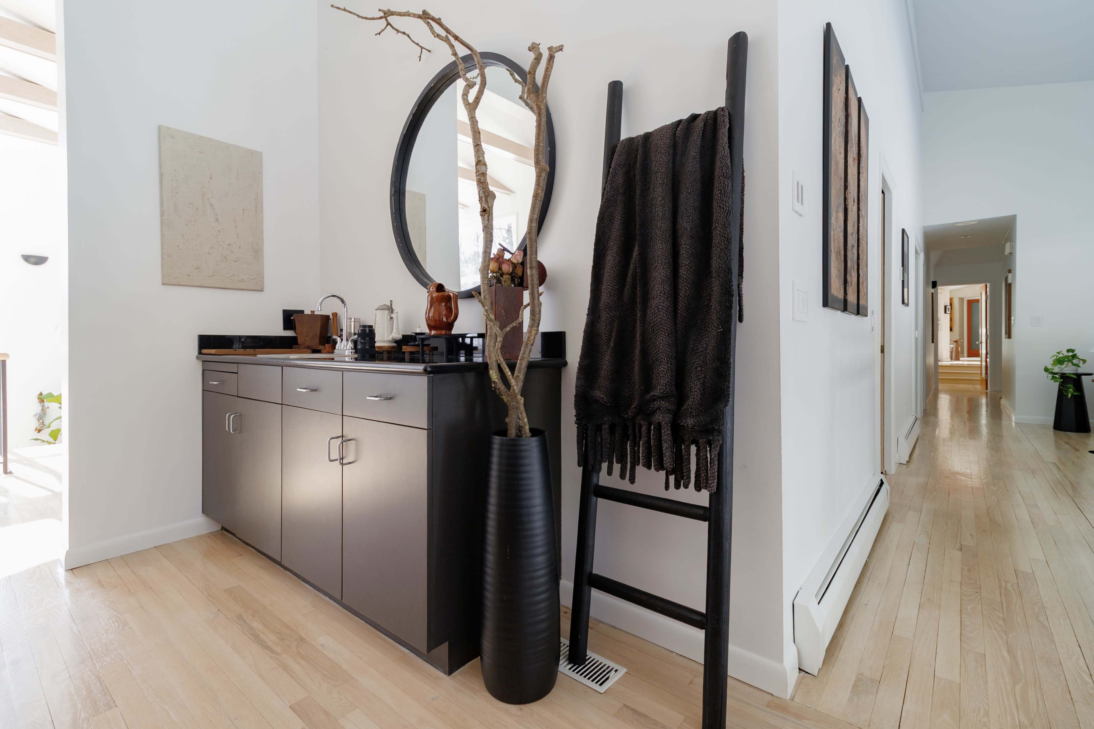 The image features a modern hallway with a sleek cabinet, a round mirror, a decorative branch arrangement, and a ladder holding a blanket.