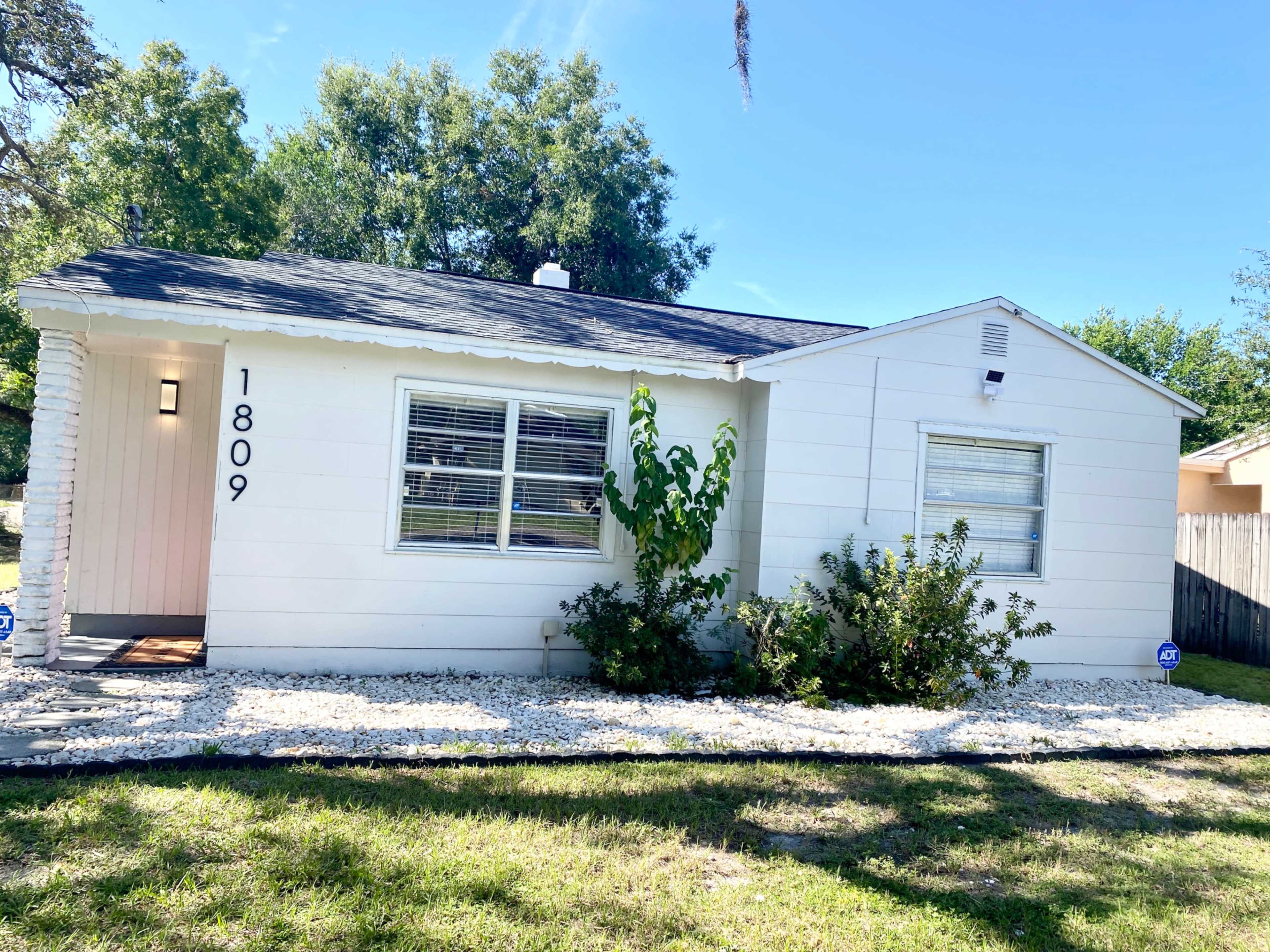 A white single-story house with a front yard and a small garden features the number "1809" displayed on the side.
