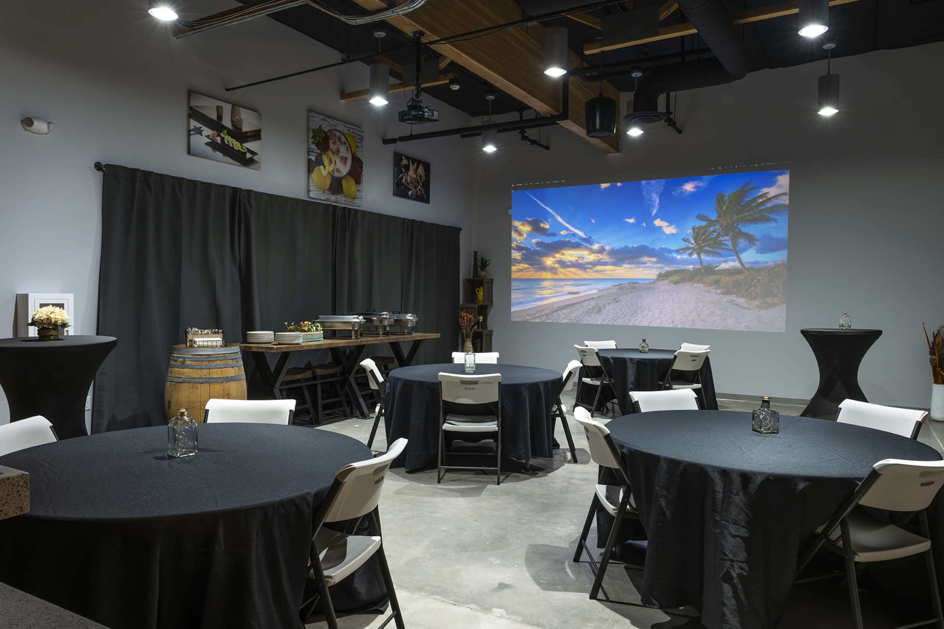 A dimly lit event space features round tables with black tablecloths, a buffet setup on one side, and a large projection of a beach scene on the opposite wall.