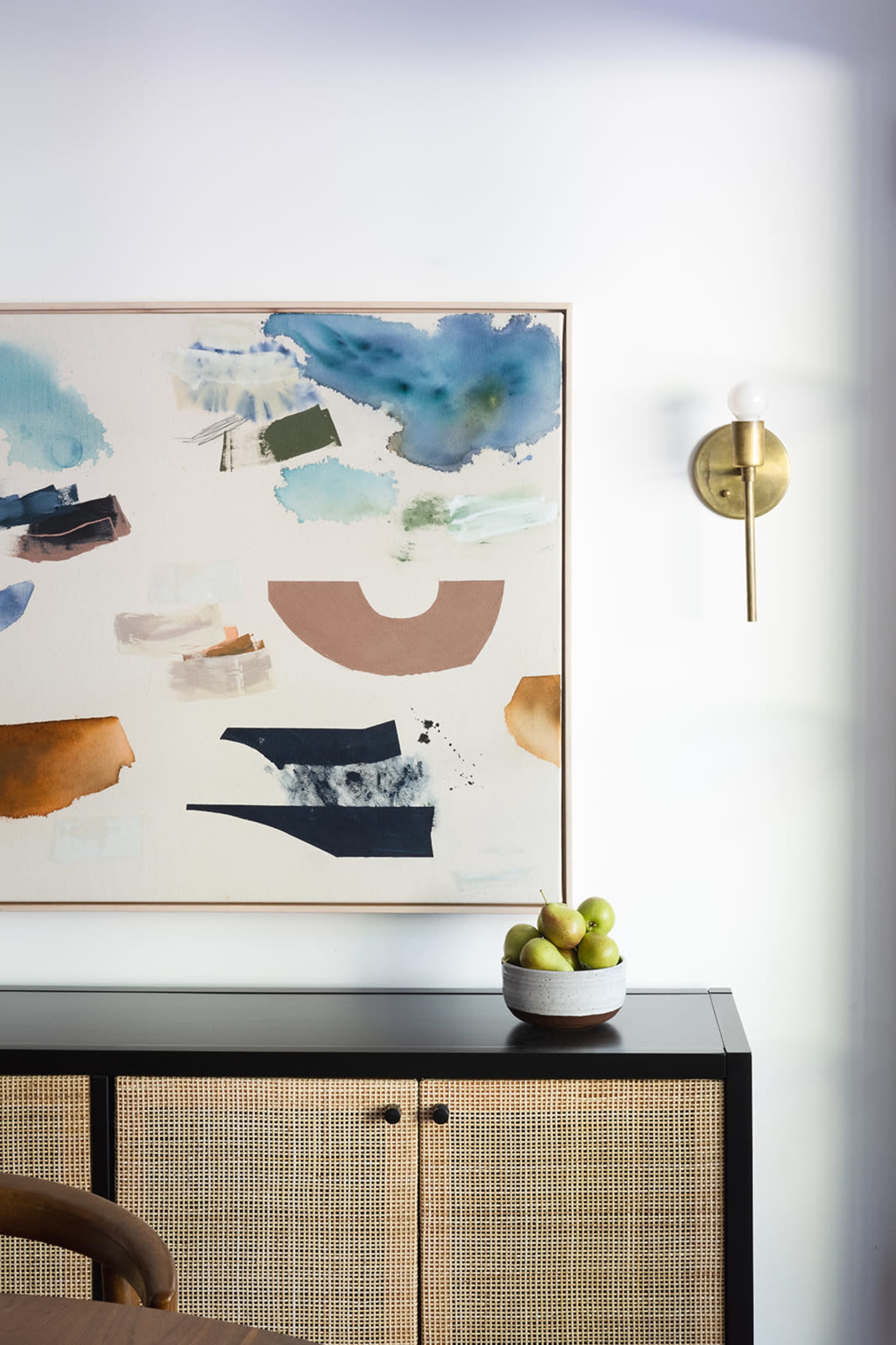 The image features a modern interior with a colorful abstract artwork hanging above a black cabinet with woven rattan doors, and a bowl of green apples on top.