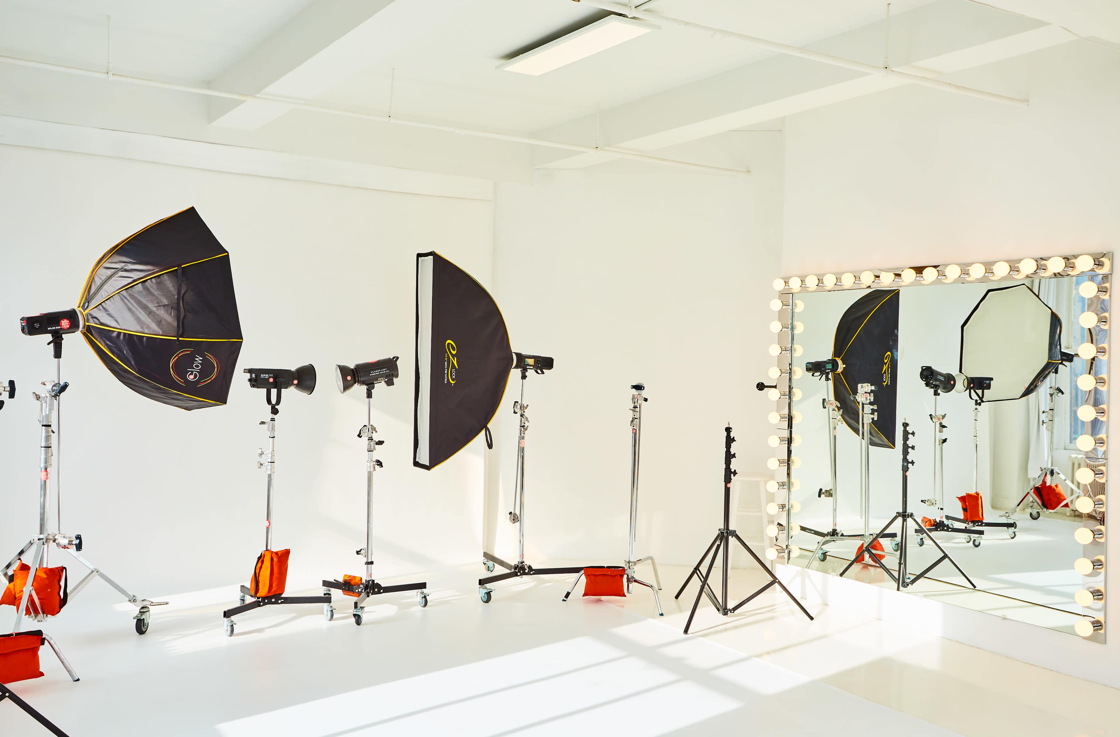 A bright photography studio features lighting equipment, including softboxes and reflectors, positioned next to a large mirrored vanity.