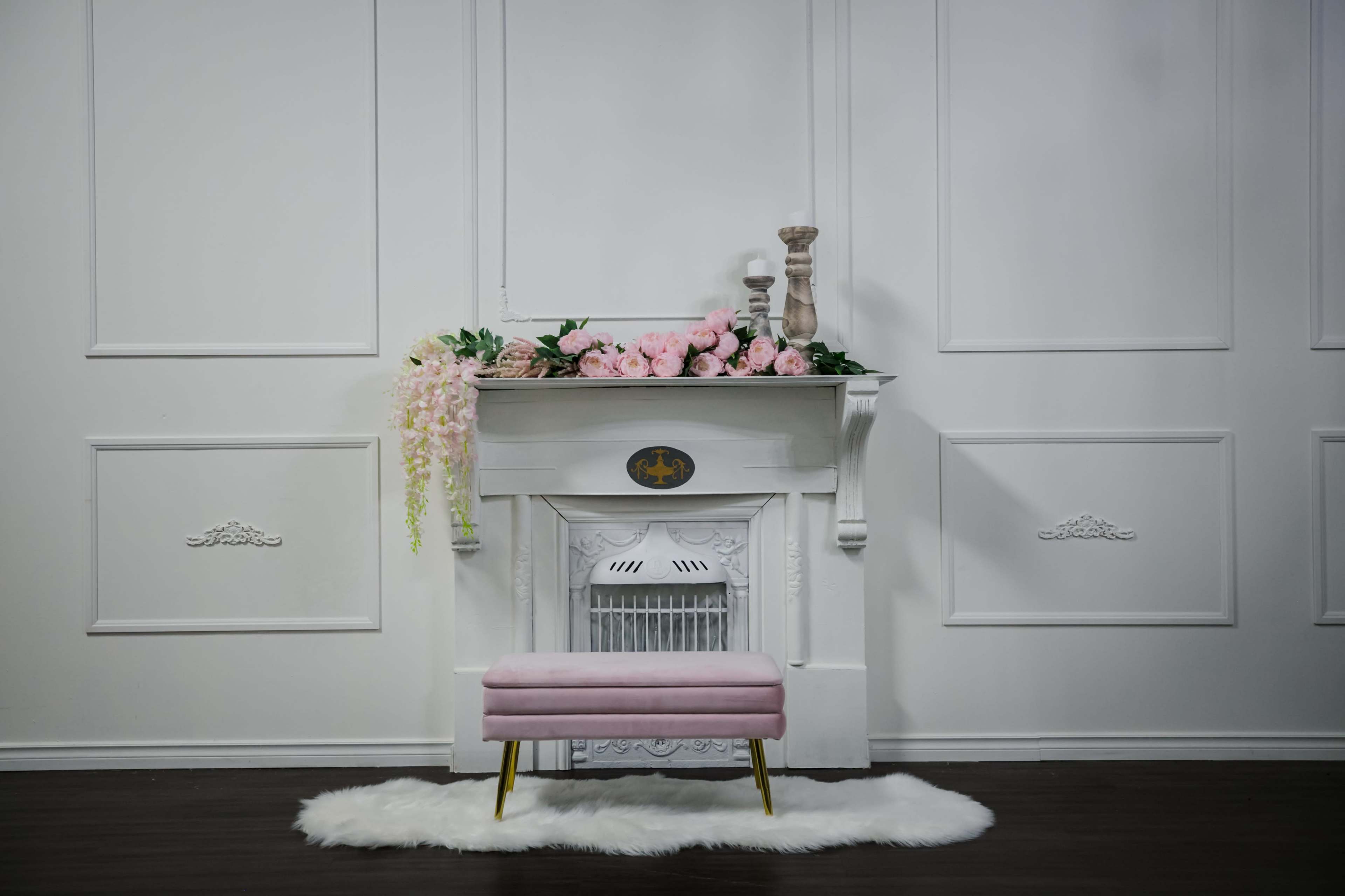 A white mantel is adorned with pink flowers and a decorative candle, with a pink bench placed on a white rug in front.