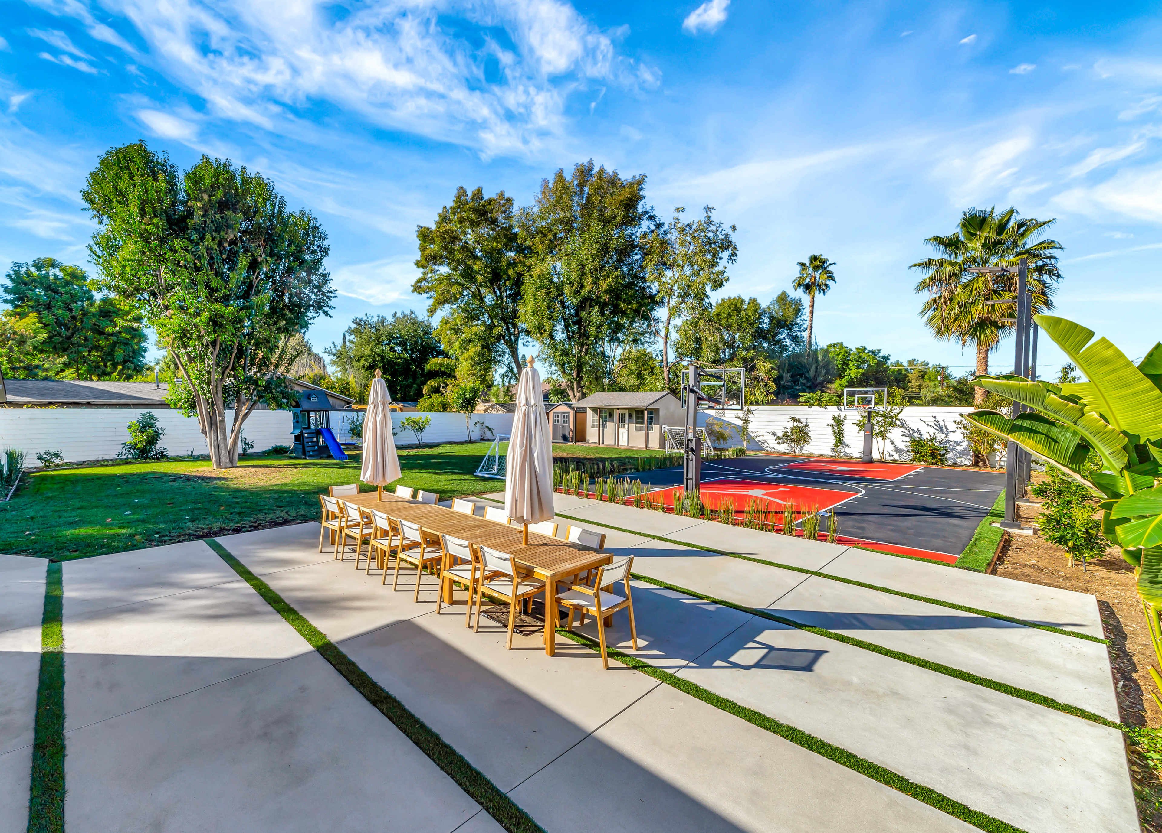 A landscaped backyard featuring a dining area with wooden tables, umbrellas, a basketball court, and a playground.