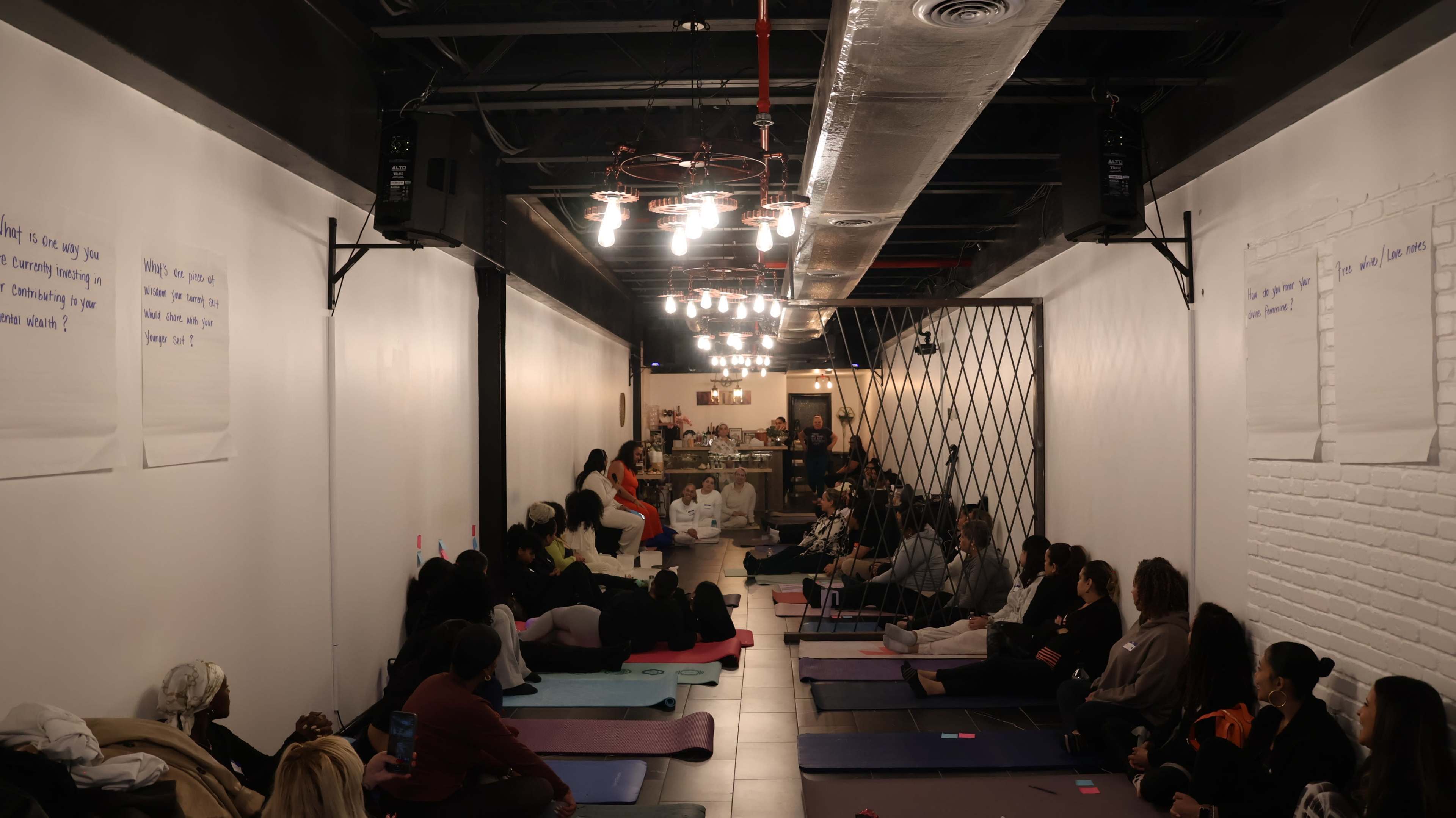A group of people is seated on yoga mats in a dimly lit room, listening to a speaker while facing a small stage area at the front.