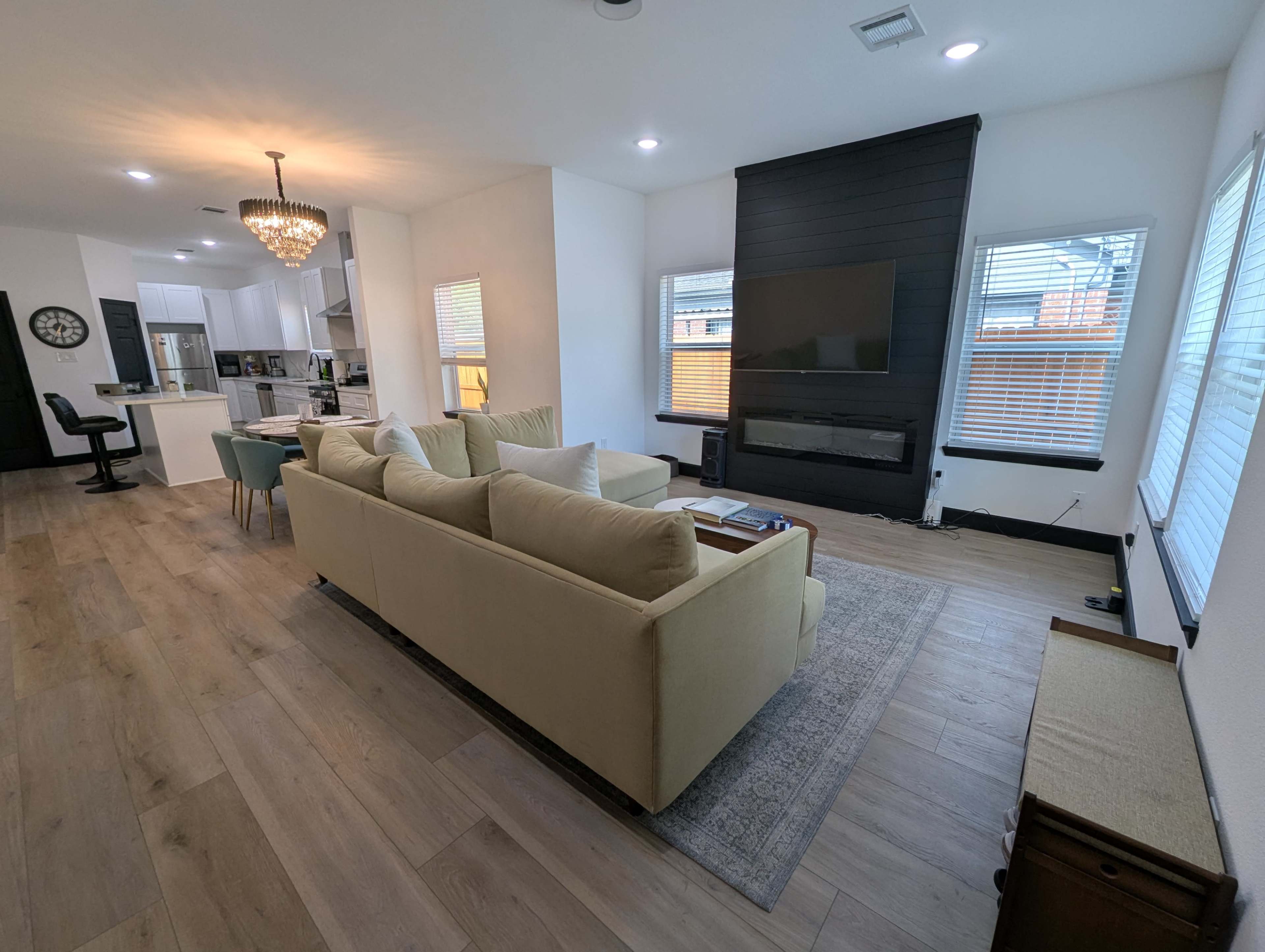 The image shows a modern living room with a beige sofa, a large TV mounted on the wall, and a dining area in the background with a chandelier and a kitchen.