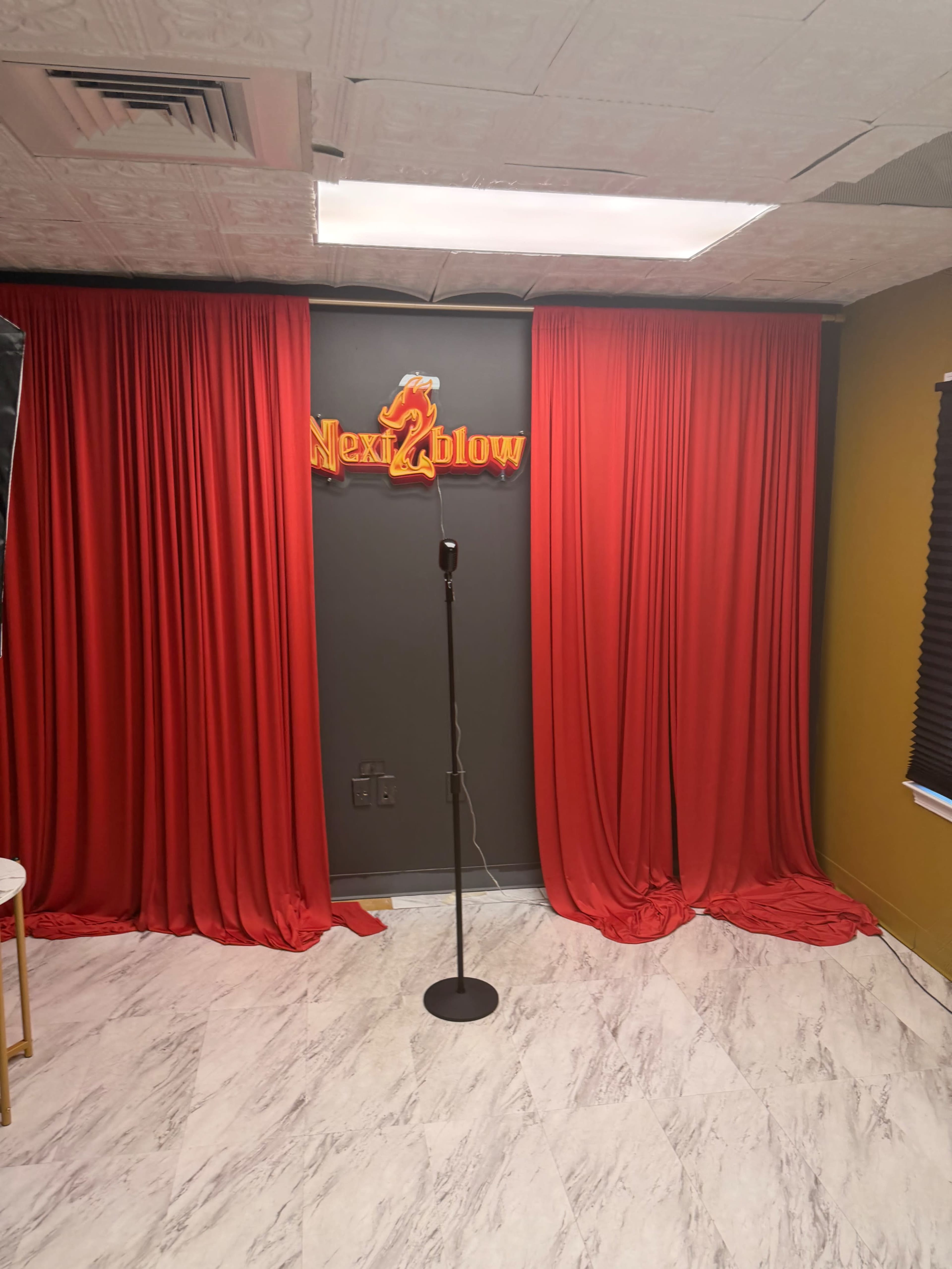 The image shows a small stage area with red curtains, a microphone stand, and a sign that reads "Next 2 Blow."
