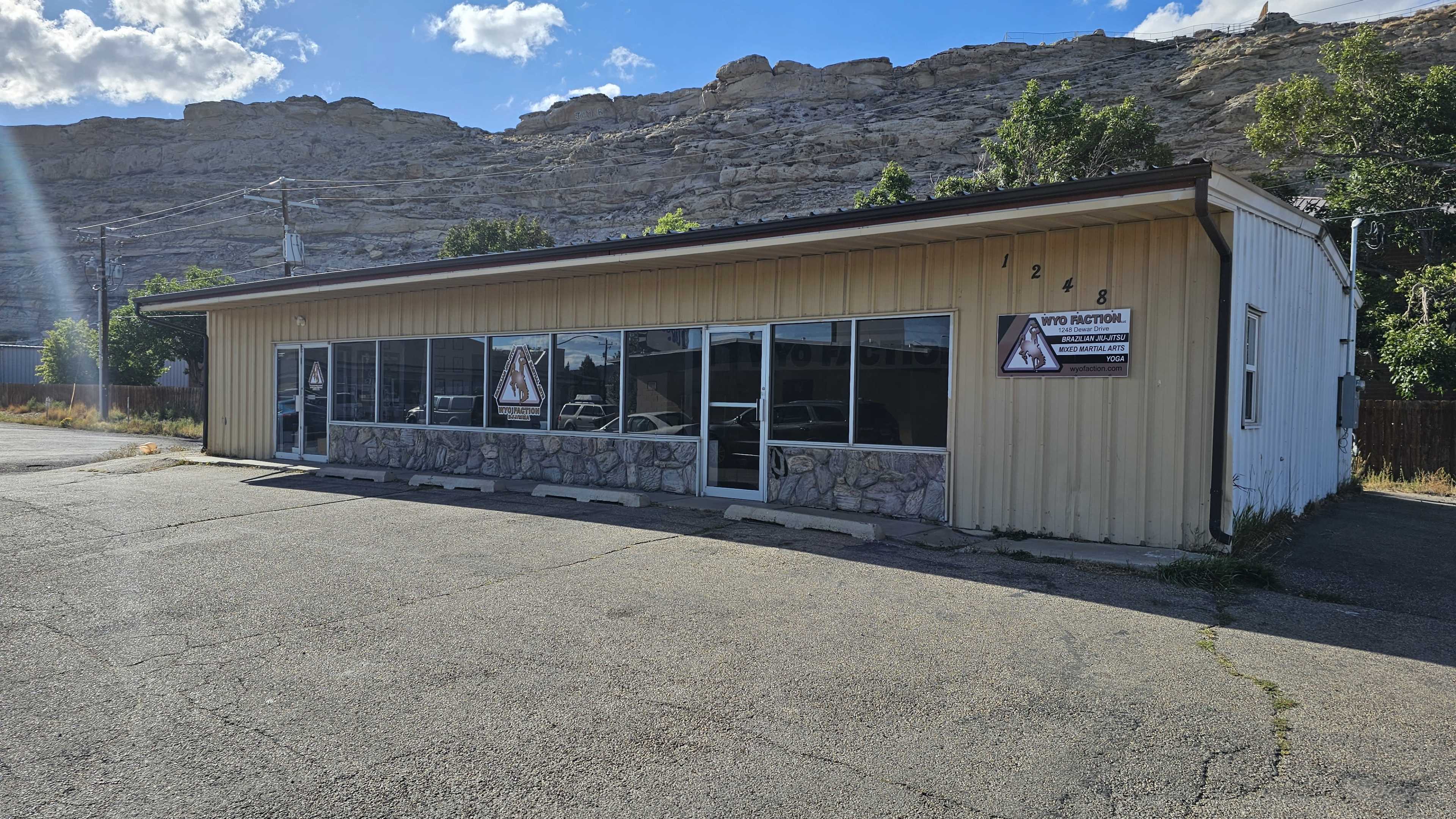 The image shows a one-story commercial building with a metal exterior, large windows, and a stone façade, situated near a rocky hillside.