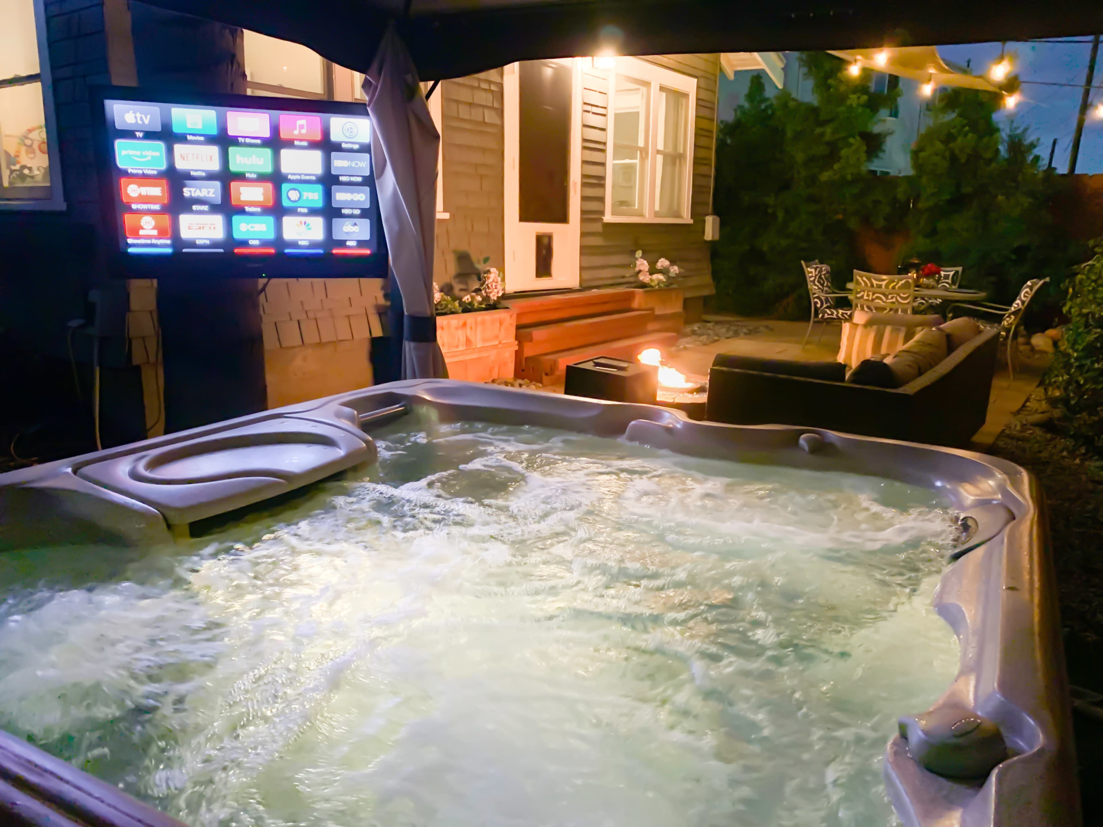 The image shows a hot tub with bubbling water under a covered patio, accompanied by a television and outdoor seating area illuminated by string lights.