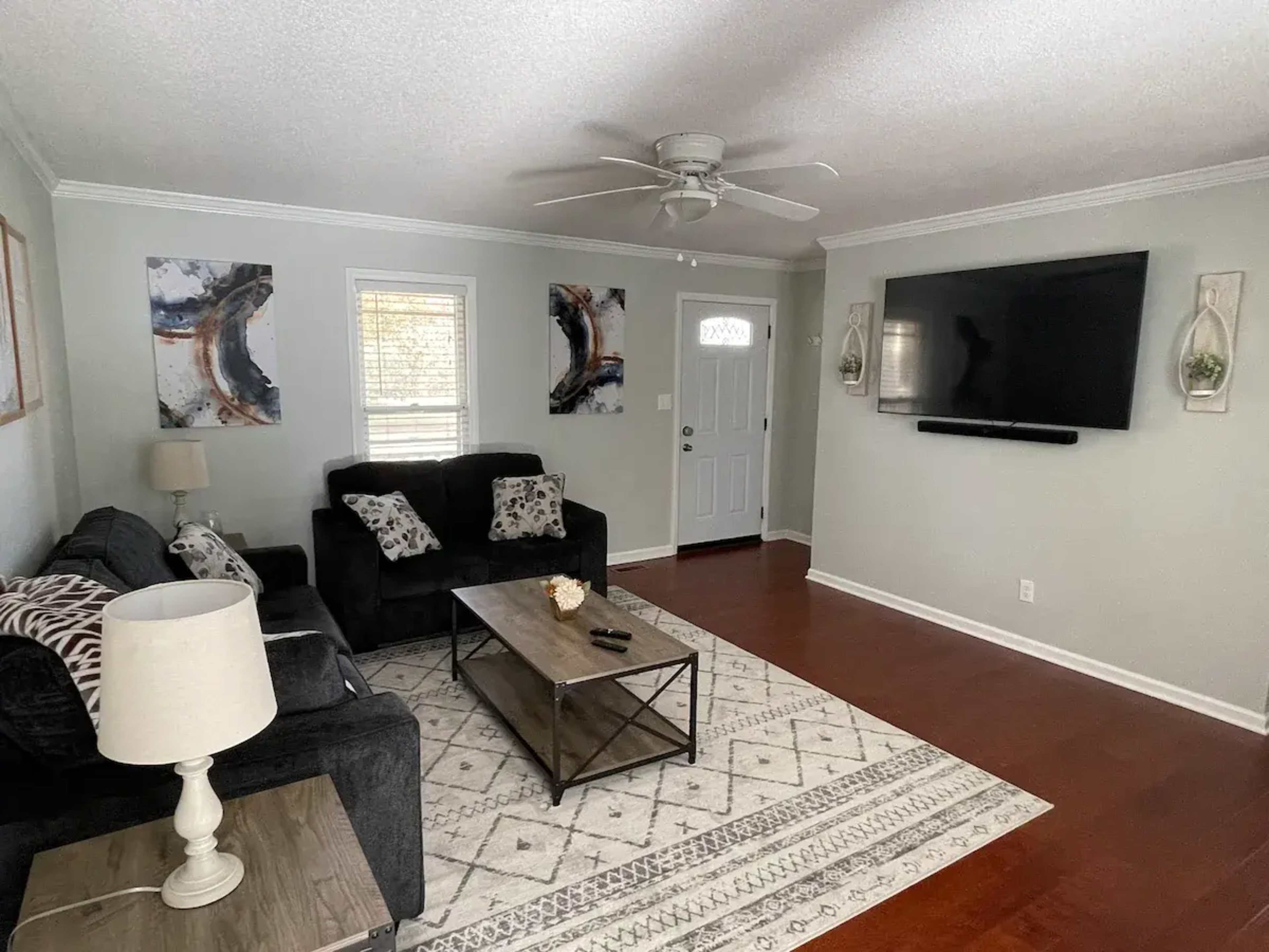 A living room with a dark sofa, a coffee table, and a mounted television on a light-colored wall, complemented by a rug and decorative wall art.