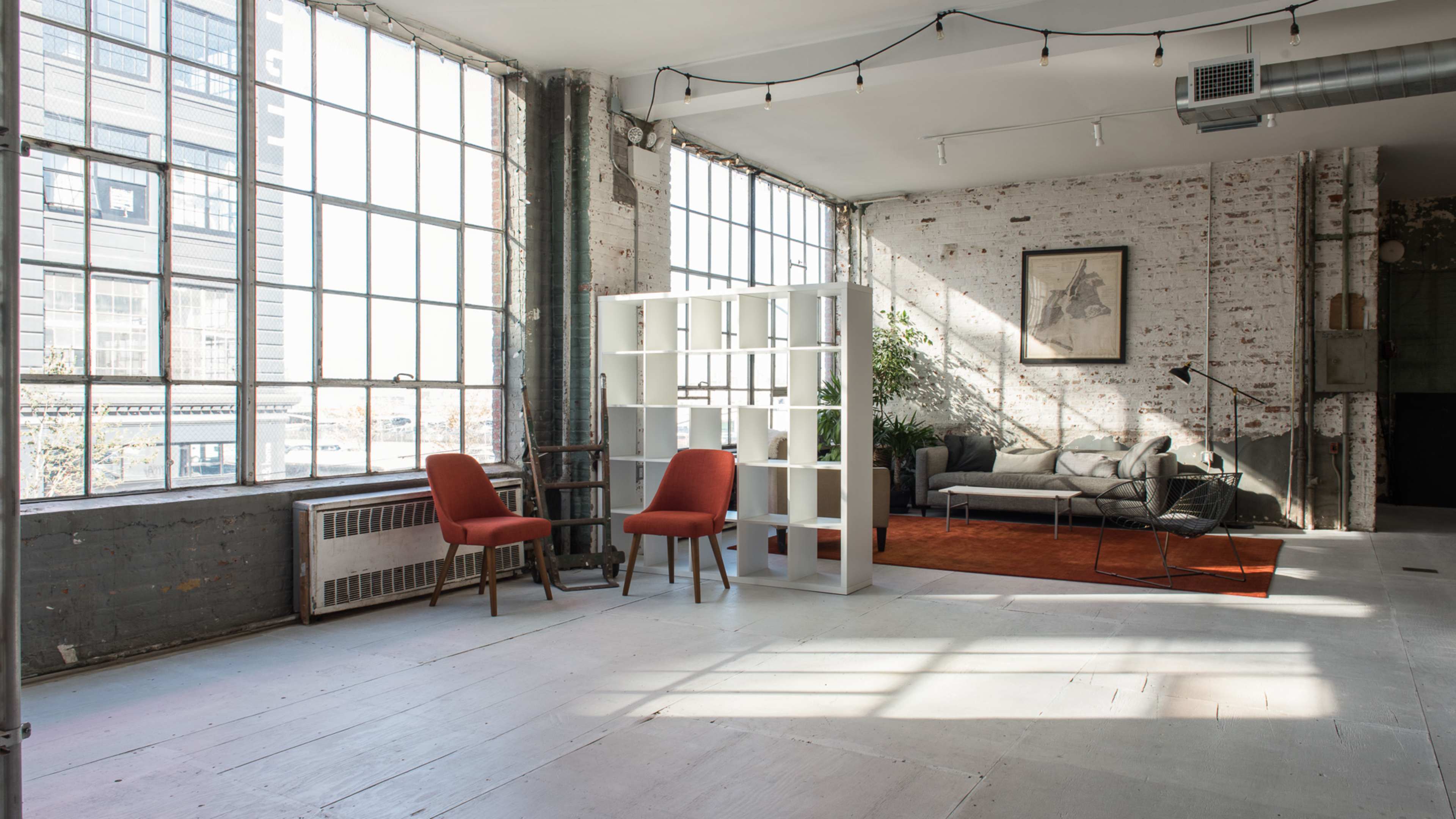 The image shows a spacious, sunlit interior with large windows, two red chairs, a white bookshelf, a gray sofa, and a potted plant on a wooden floor.