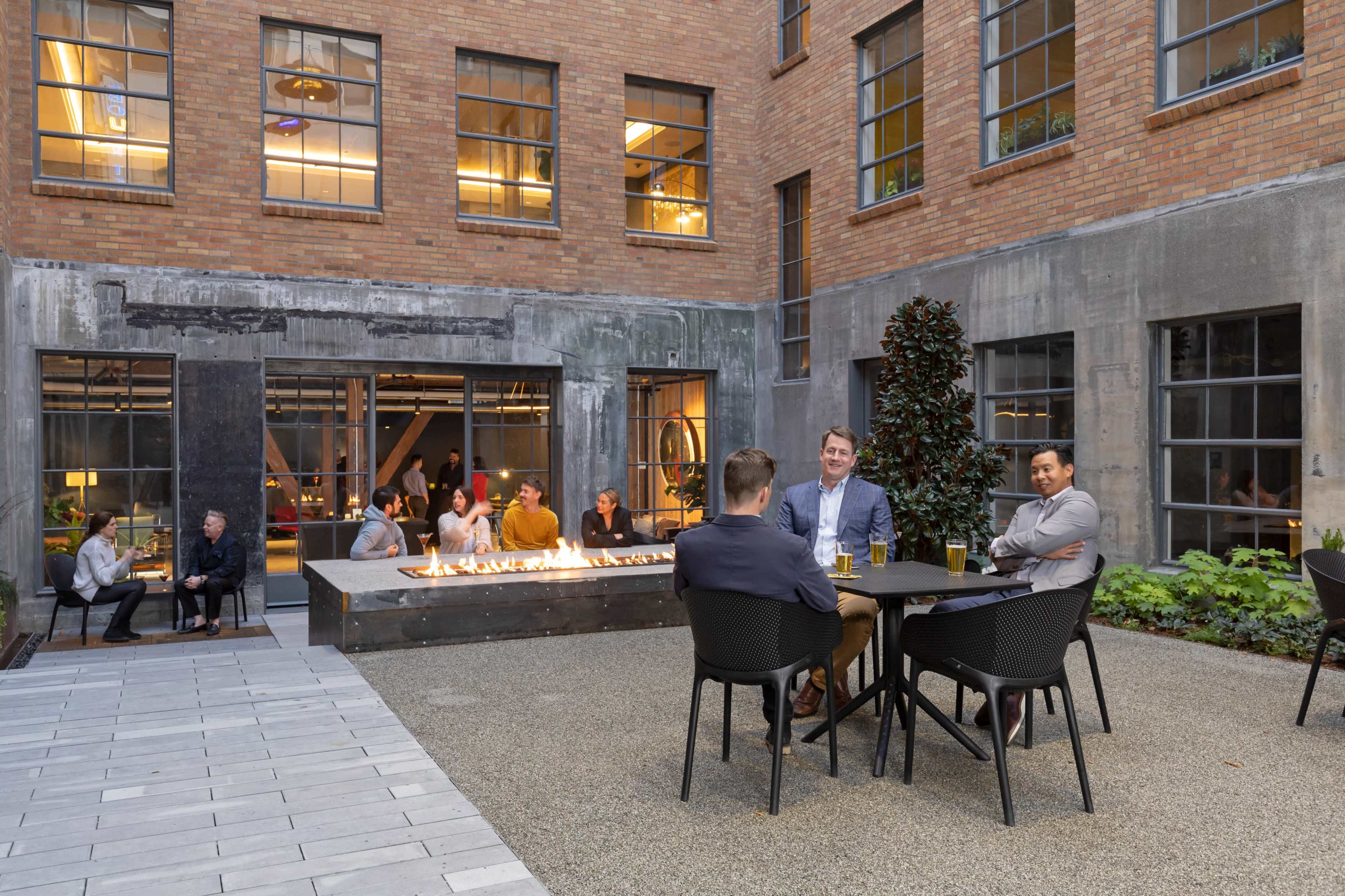 A group of people sits at tables around a fire pit in a courtyard surrounded by brick walls and large windows.