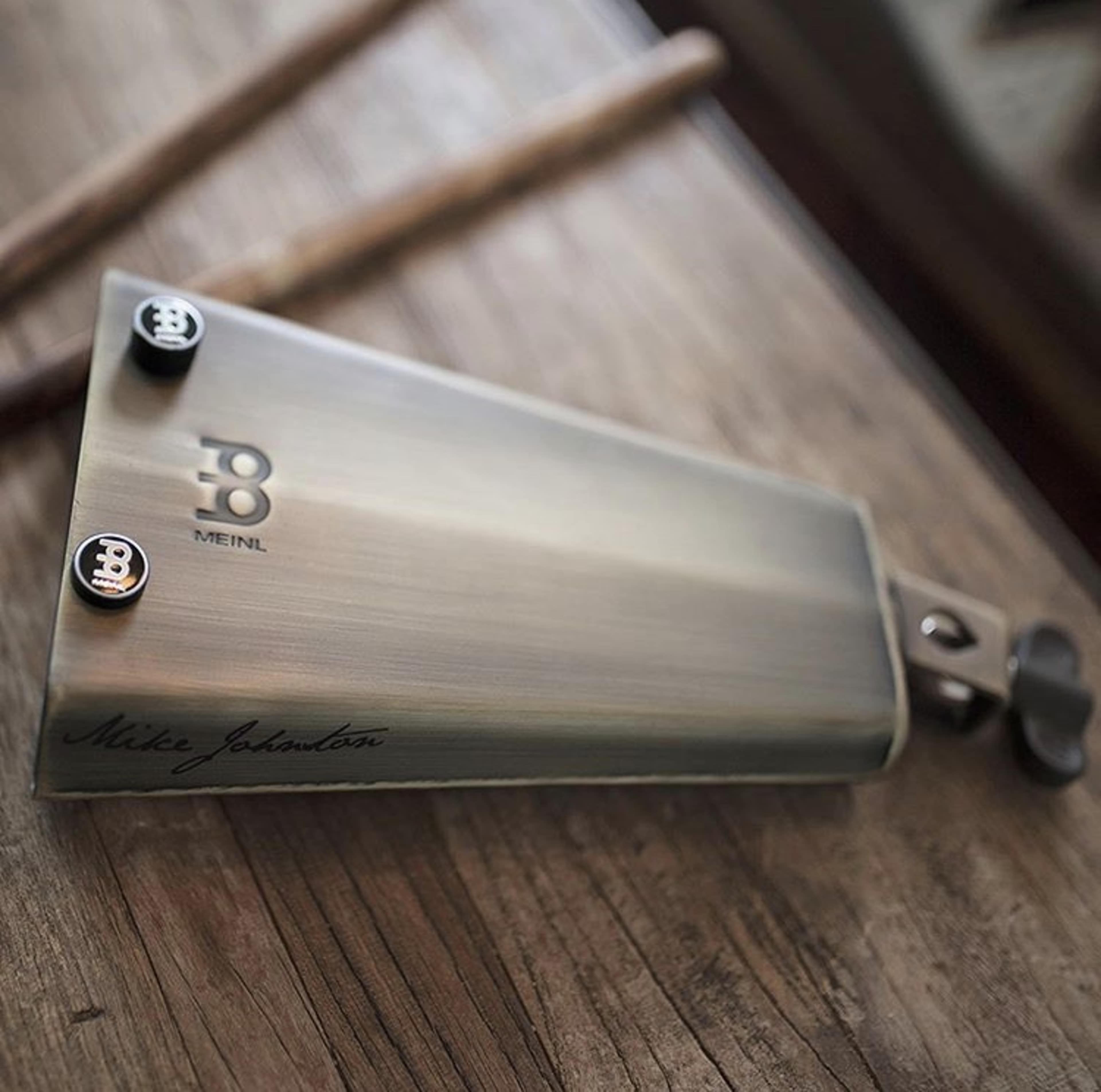 A Meinl cowbell with the signature "Mike Johnston" is positioned on a wooden surface next to drumsticks.