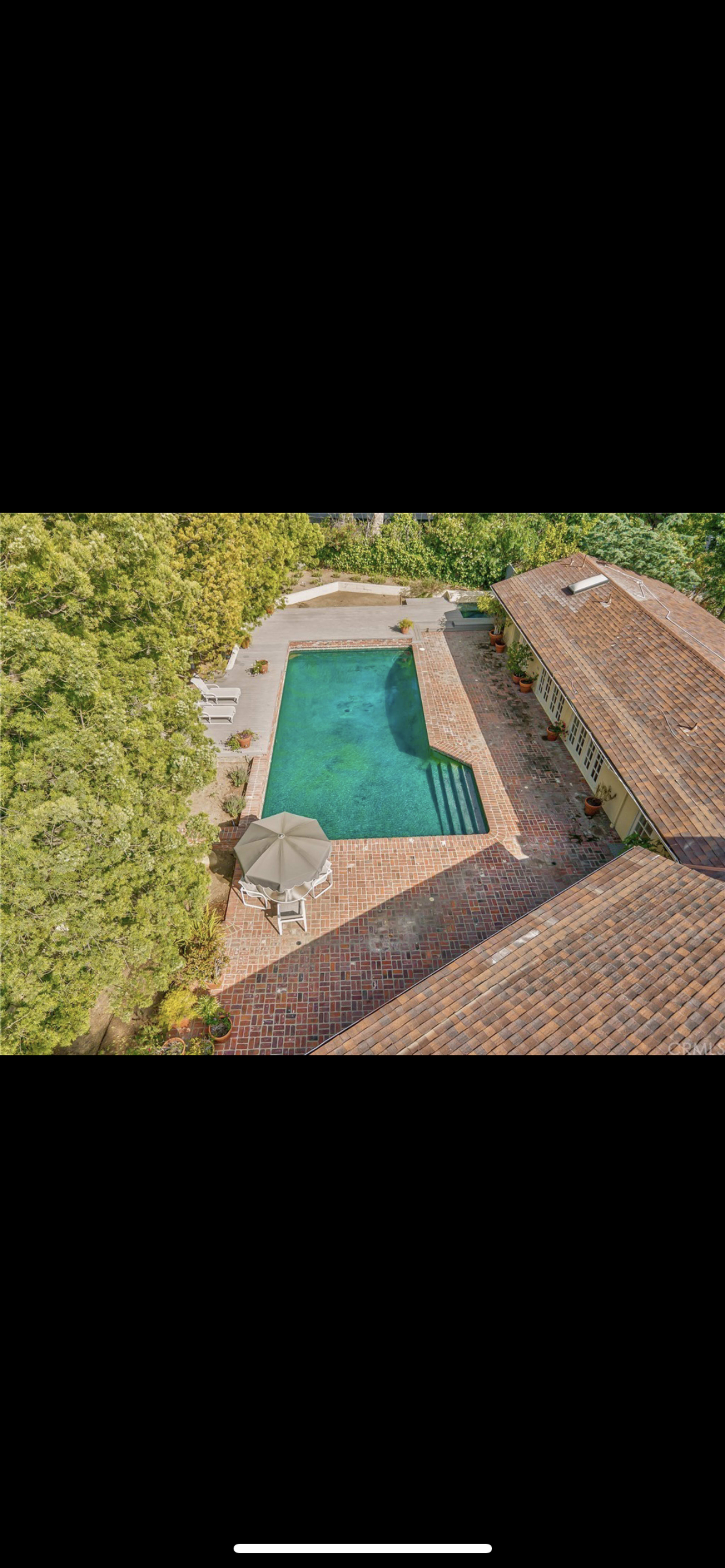 An aerial view shows a rectangular swimming pool surrounded by a brick patio and a shaded umbrella area, adjacent to a house with a tiled roof.