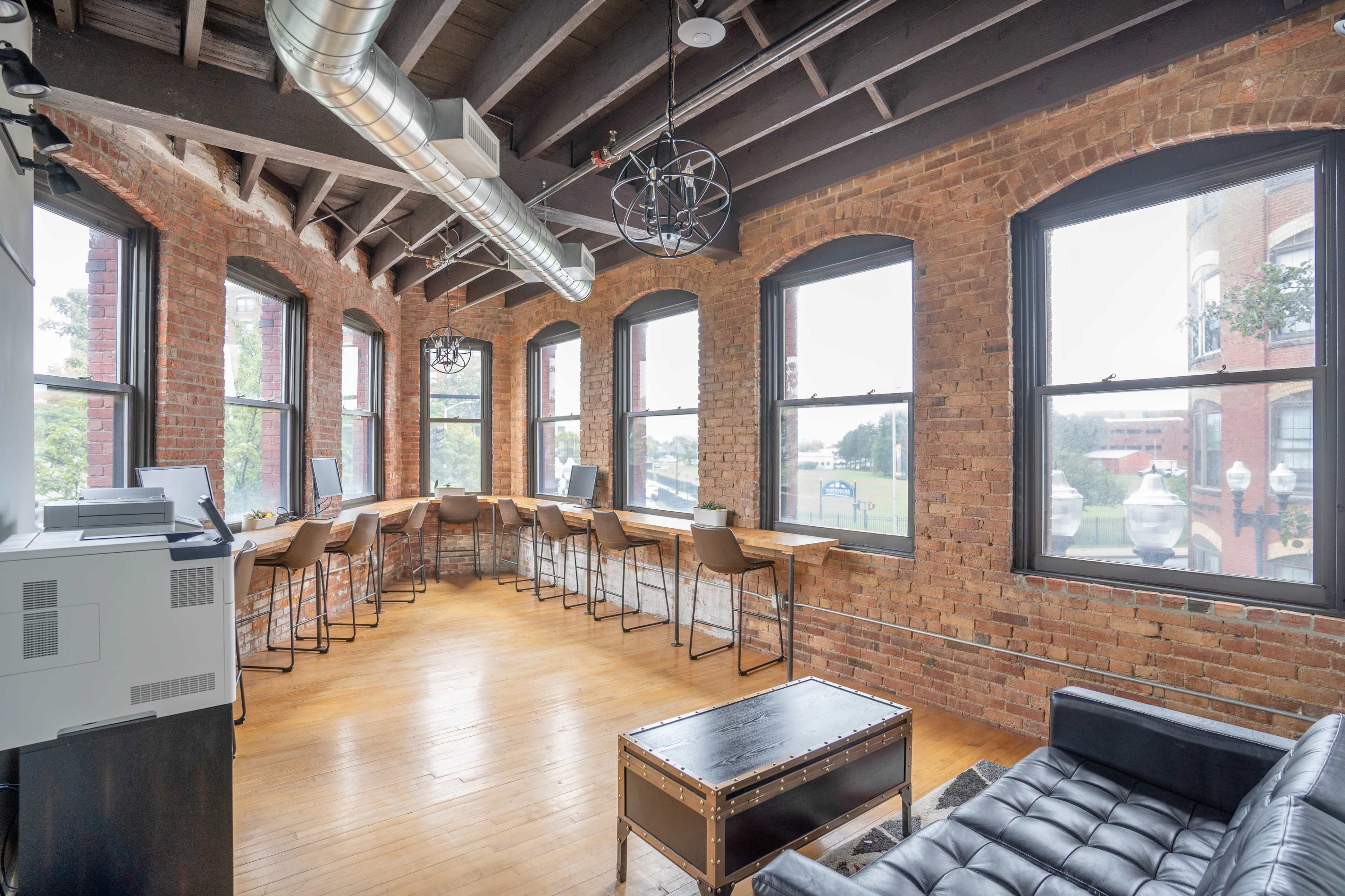 The image shows a modern office space with exposed brick walls, large windows, and wooden flooring, featuring a long wooden table with high chairs along one side.