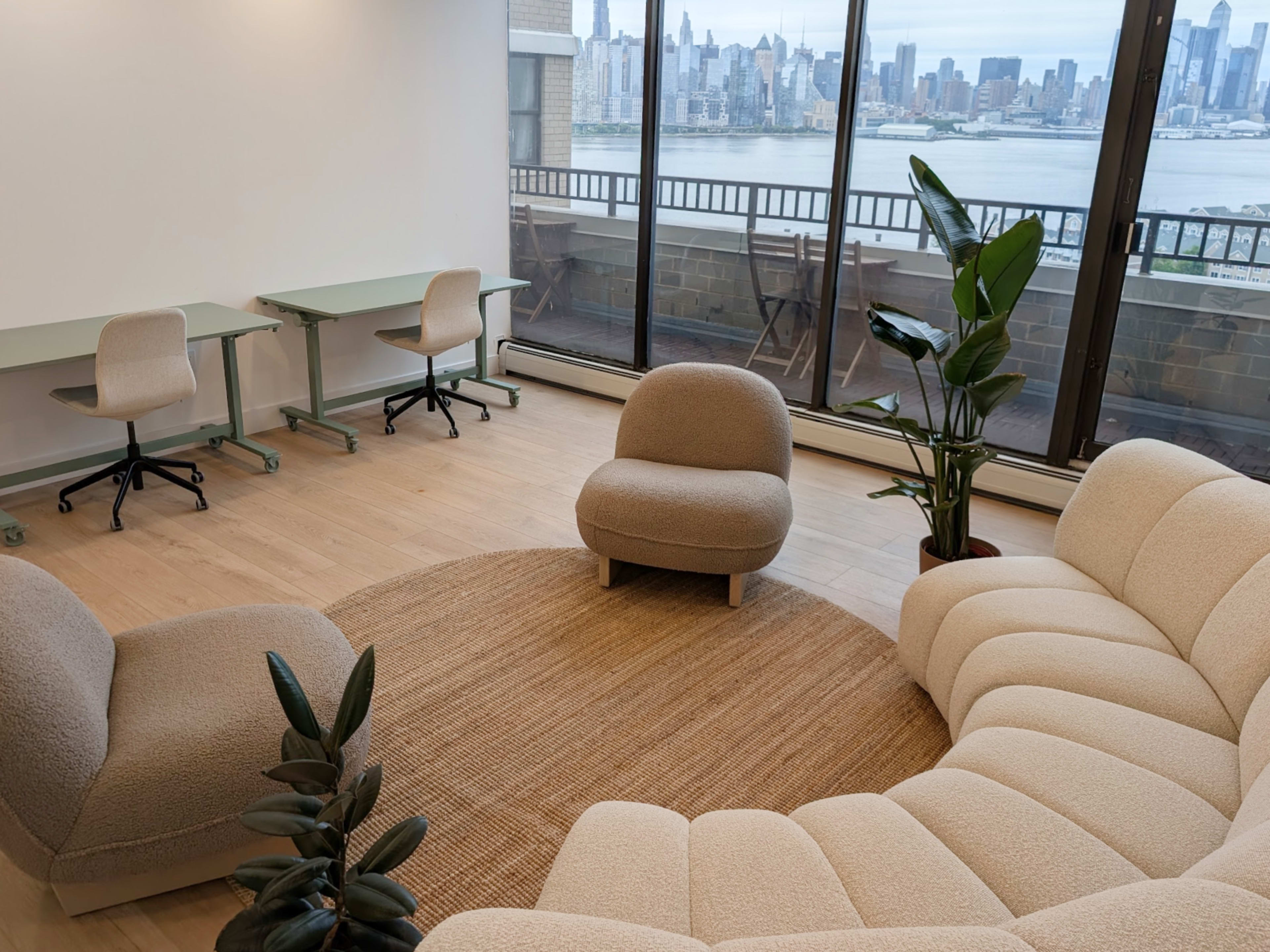 A cozy seating area featuring a curved sofa and armchair, with a view of a city skyline through large glass windows.