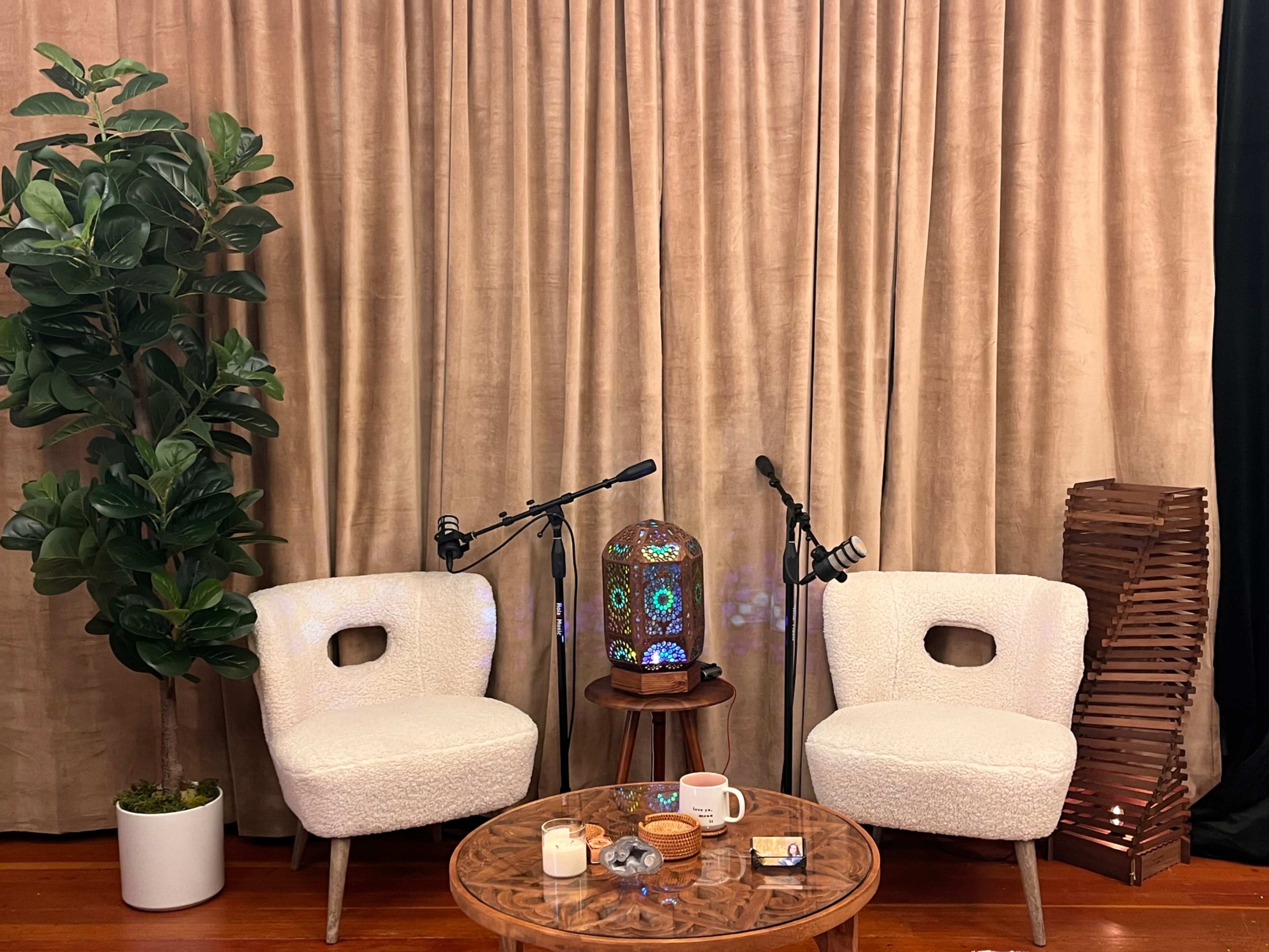 The image shows two cozy chairs with microphones set up beside a small table, in front of a large curtain and a decorative lamp.