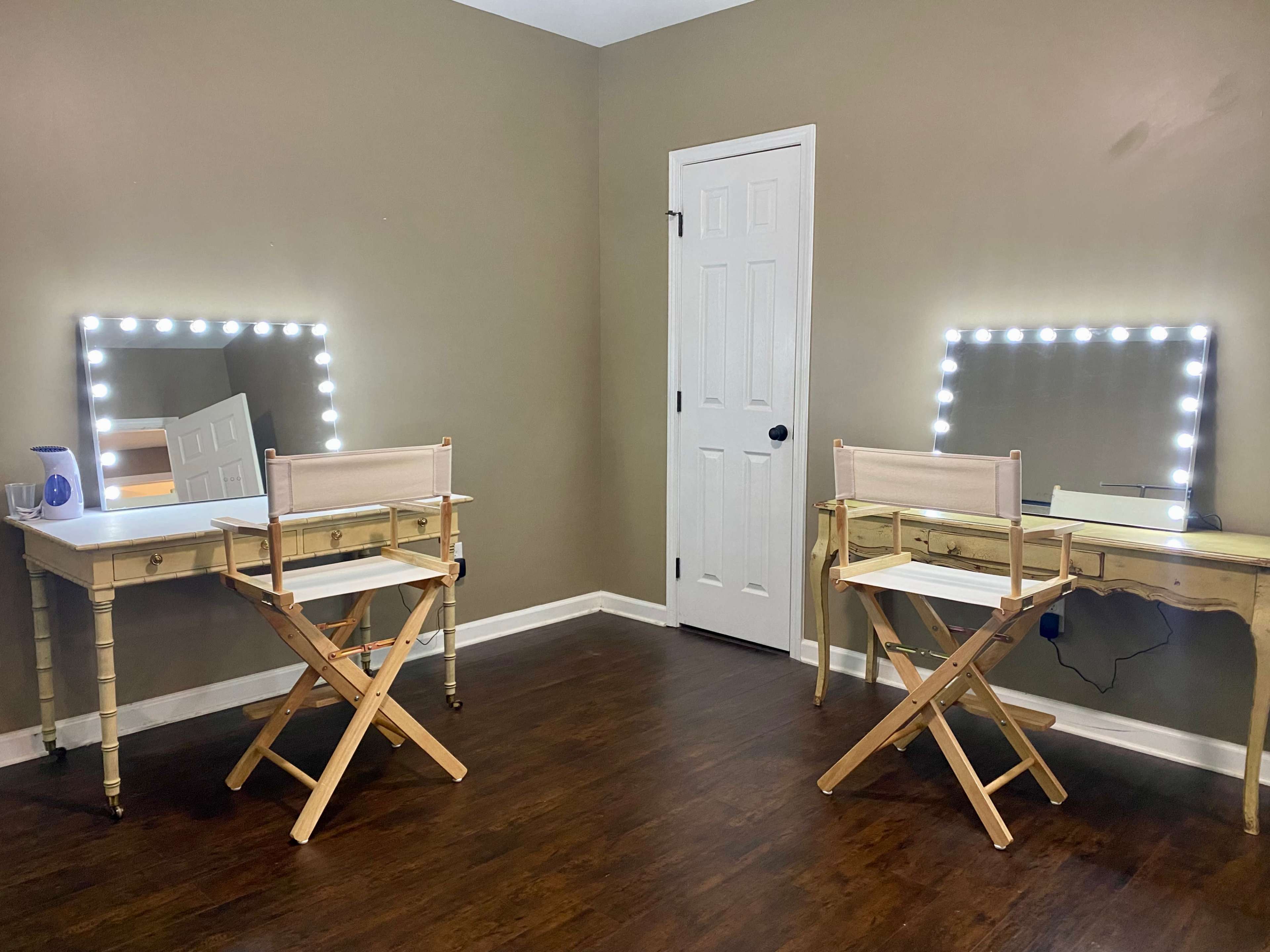 The image shows a room with two lighted vanity mirrors and folding chairs placed on opposite sides of a beige wall.