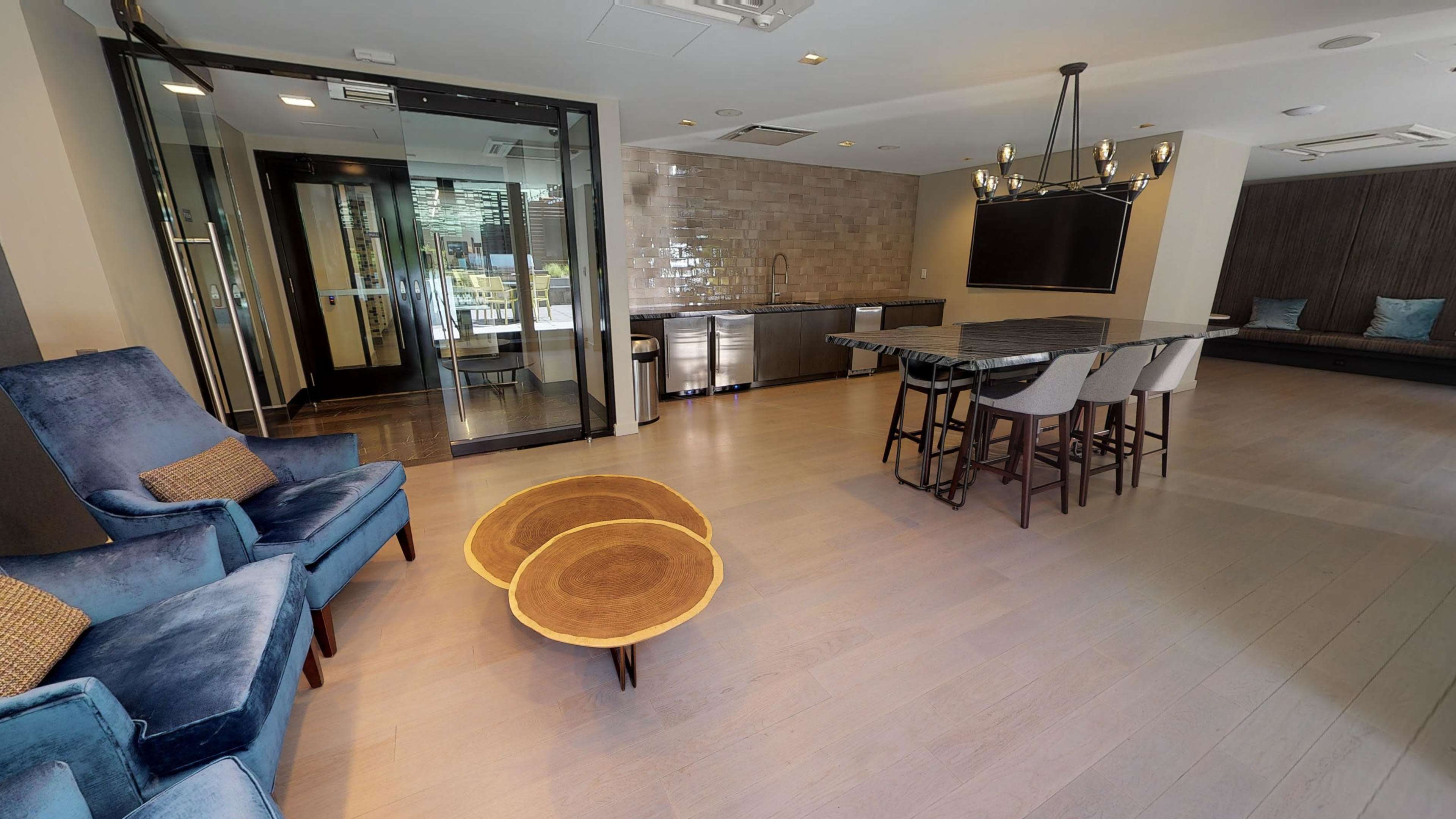 The image shows a modern lounge area with two blue chairs, a wooden table, and a kitchen space featuring stainless steel appliances and a large television on the wall.