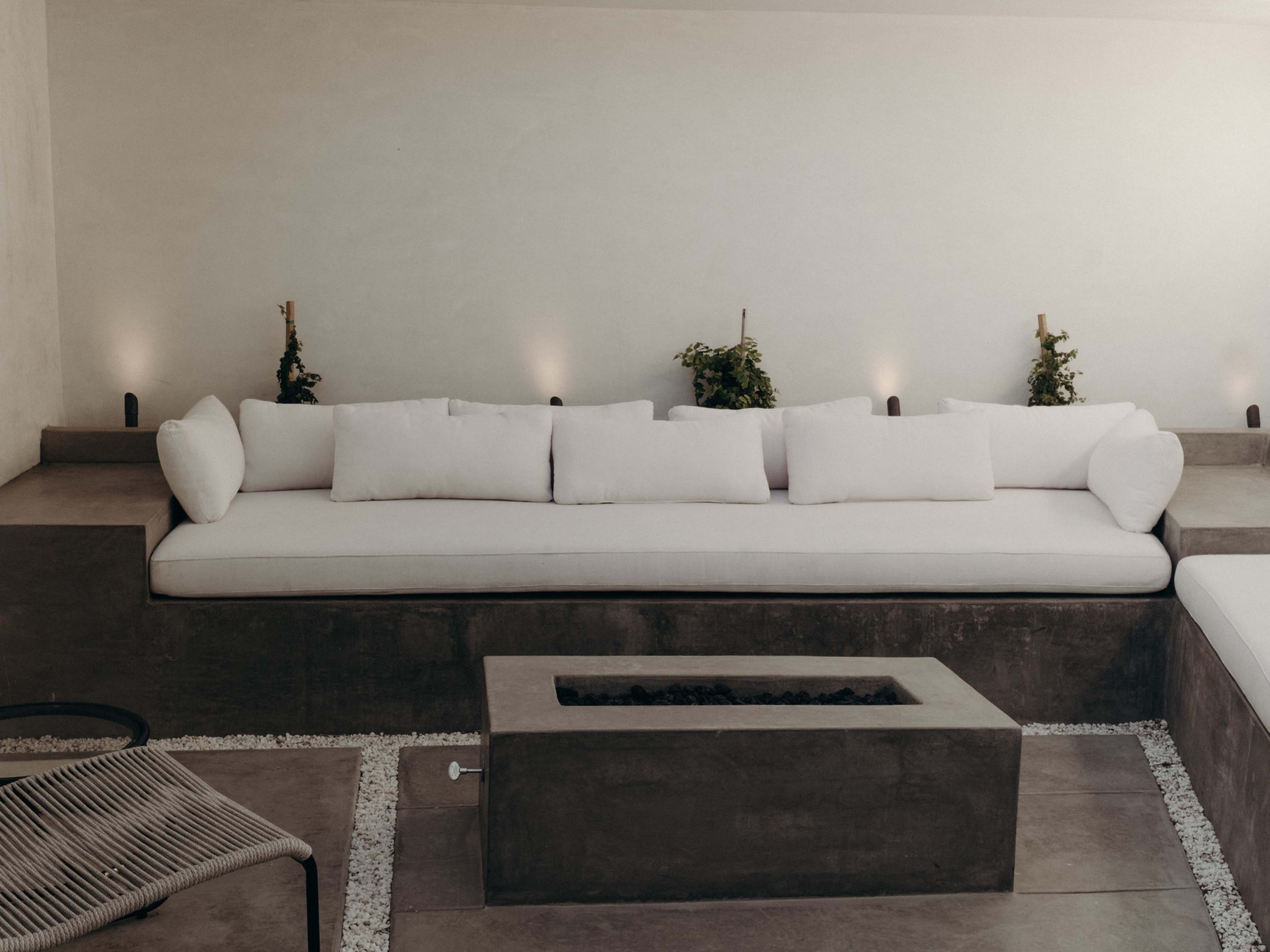A minimalist outdoor seating area with a long, cushioned bench against a plain wall, accompanied by a stone fire pit and small potted plants.