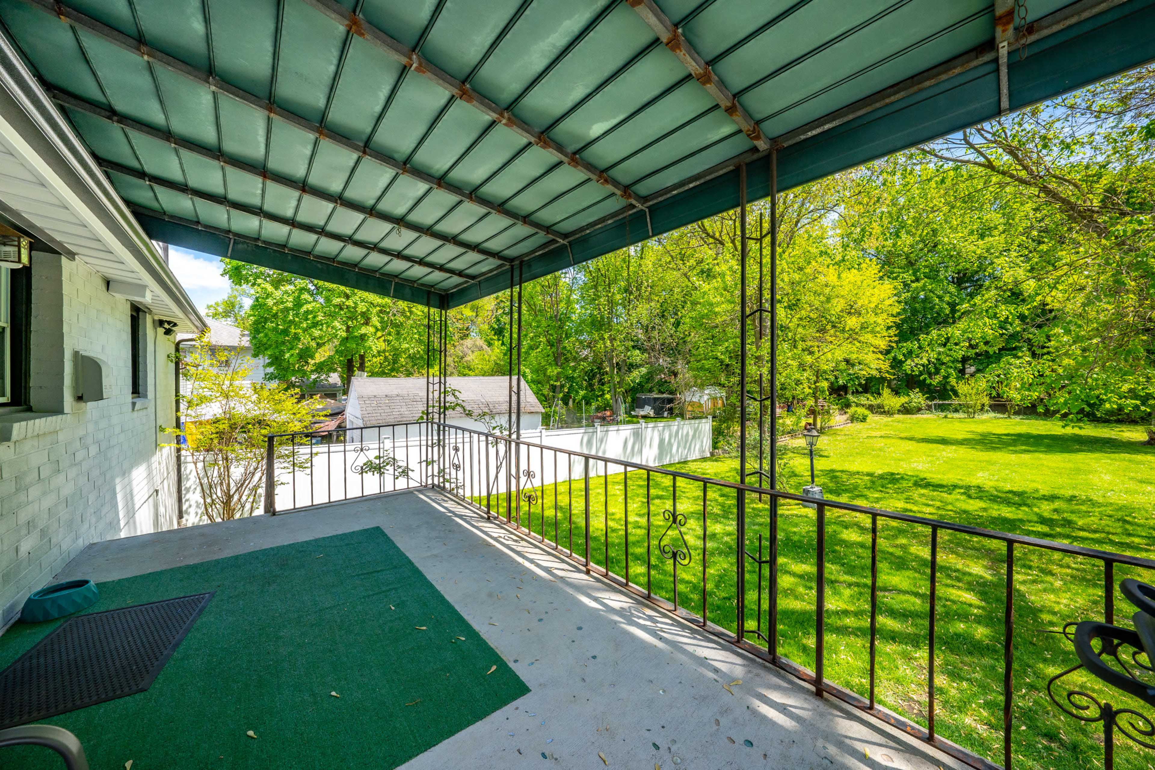 The image shows a covered porch with a green mat, overlooking a backyard filled with trees and a grassy area.