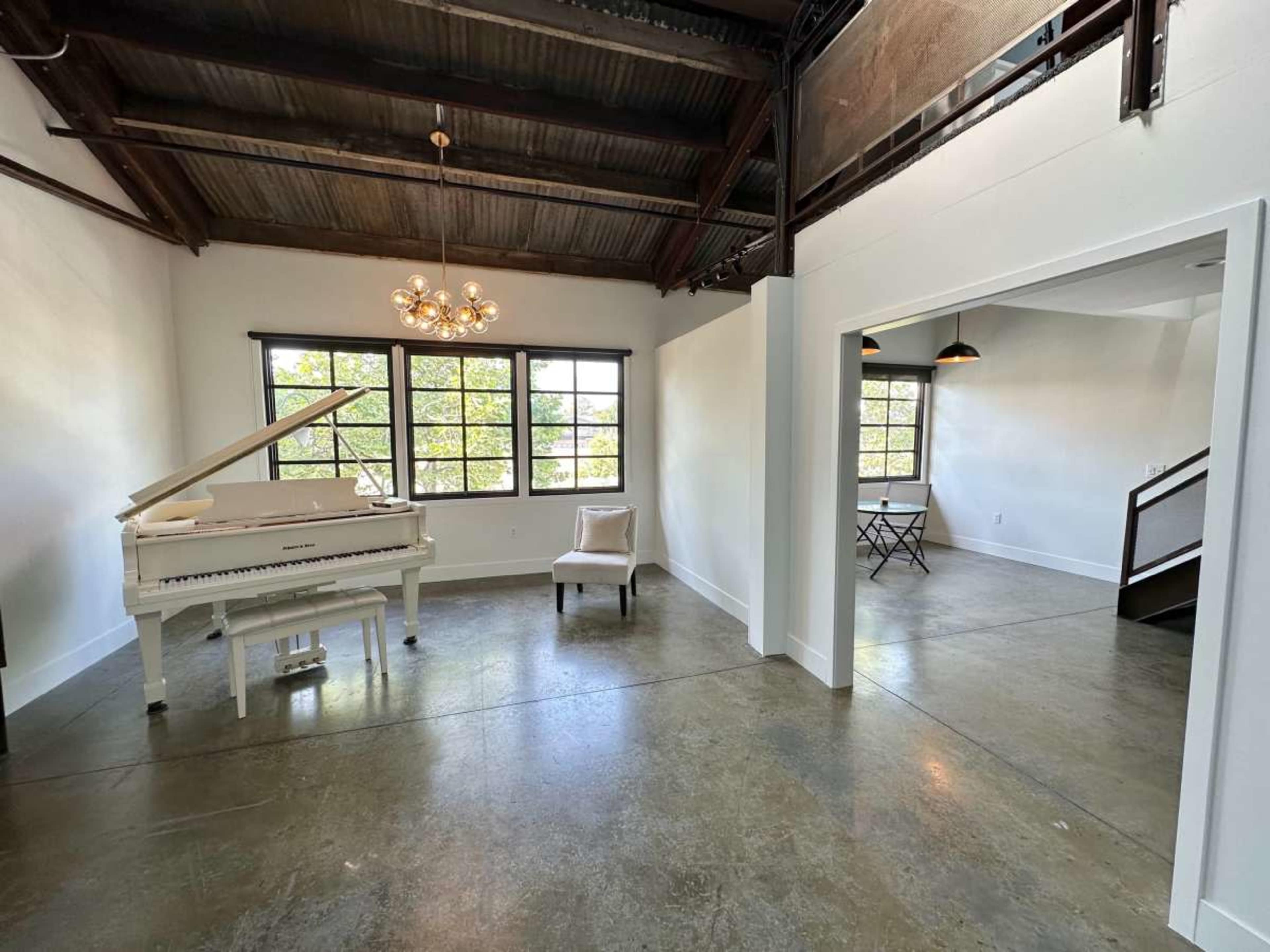 The image shows a spacious room with high ceilings, featuring a white grand piano, a chair, and a small table near large windows.