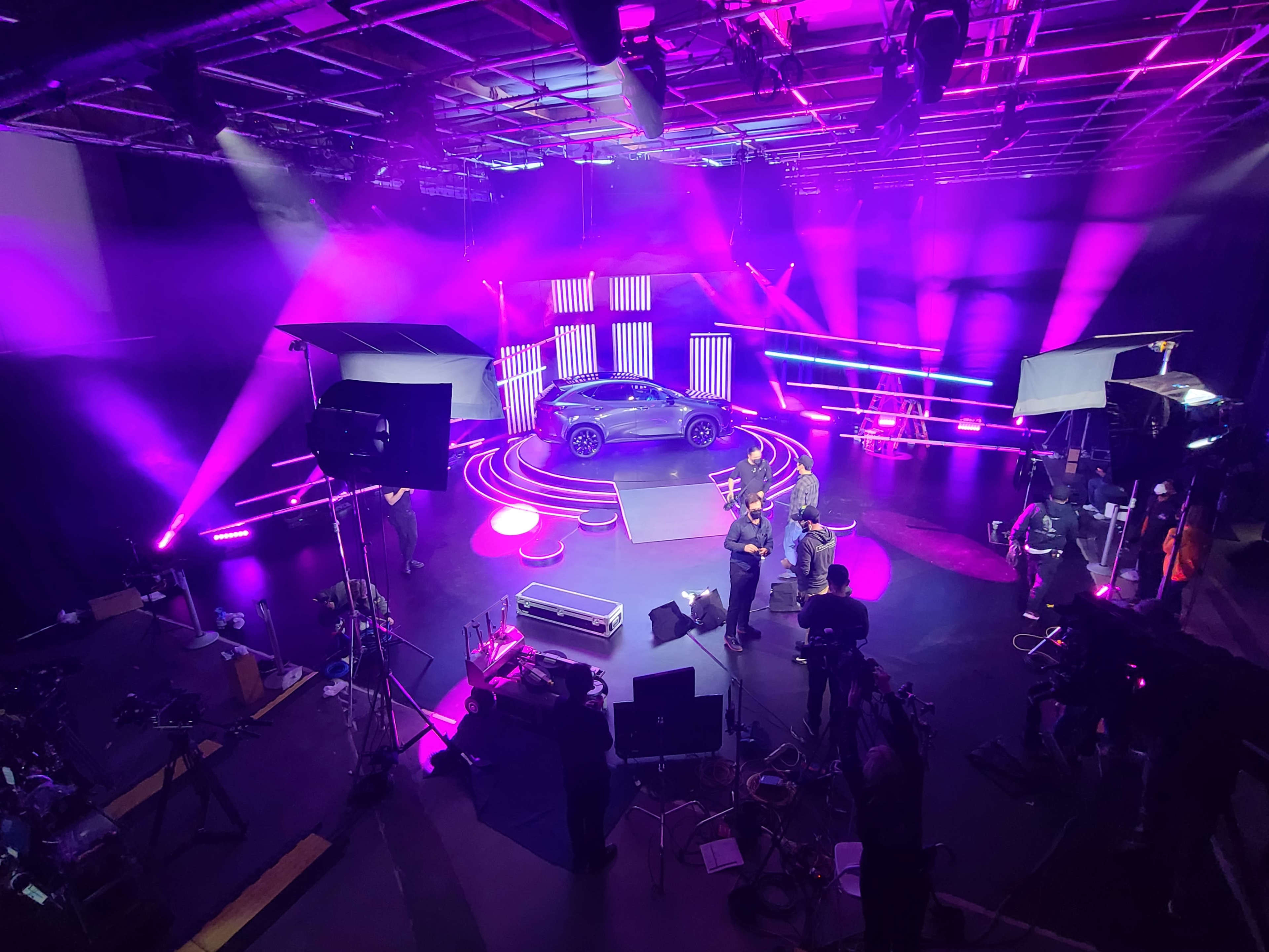 A sleek car is showcased on a circular platform surrounded by vibrant lights and film equipment in a studio setting.