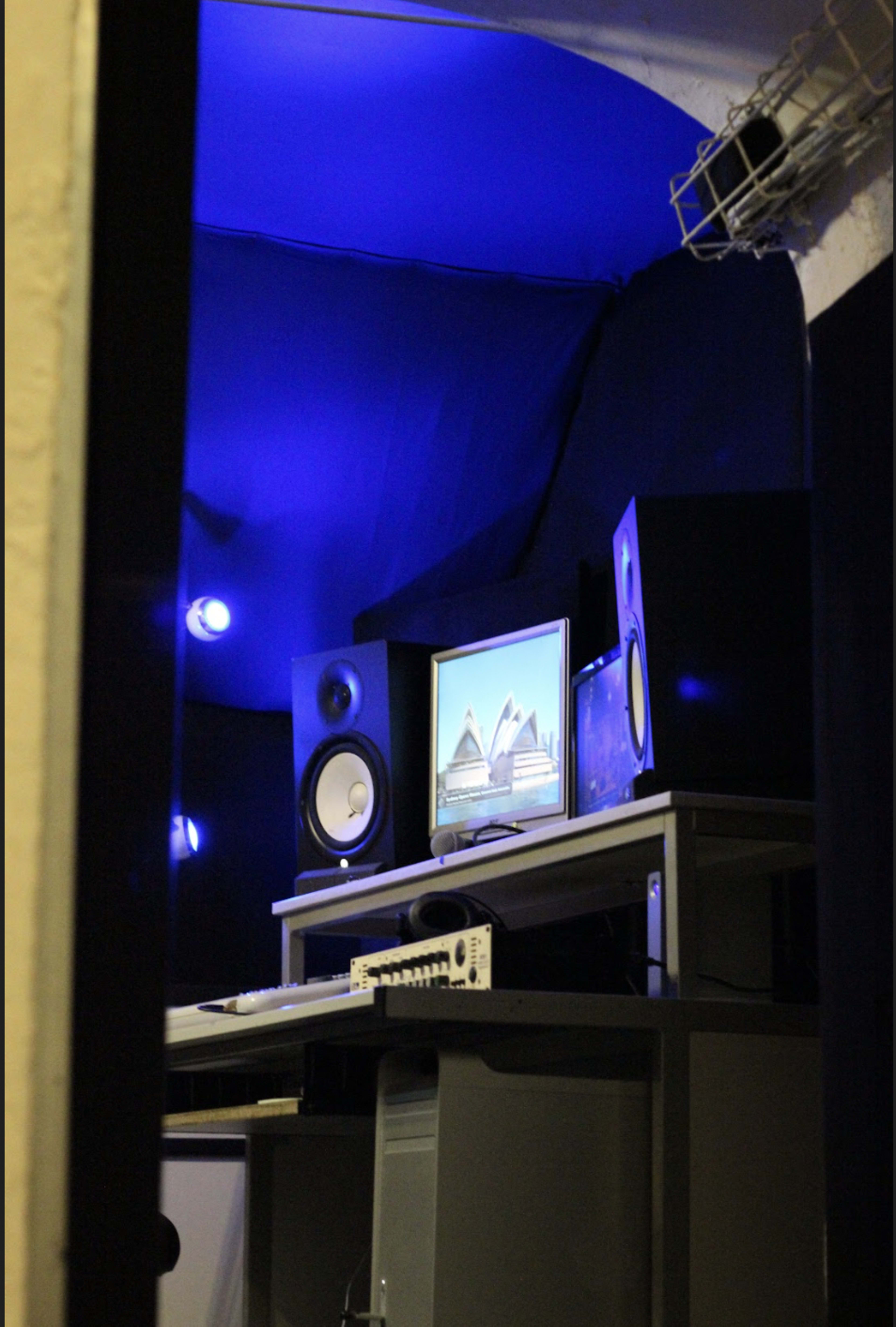 The image shows a music production studio with a computer monitor, audio speakers, and blue ambient lighting.