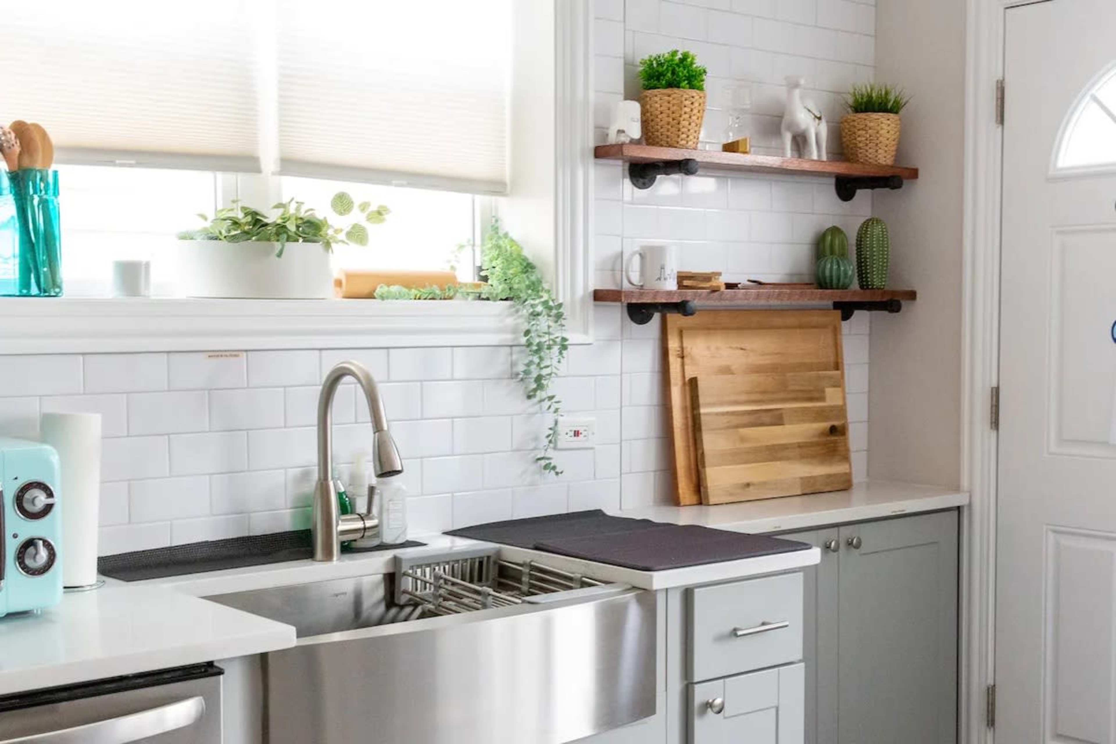A modern kitchen features a stainless steel sink, white tiled walls, and wooden shelves holding decorative plants and kitchen items.