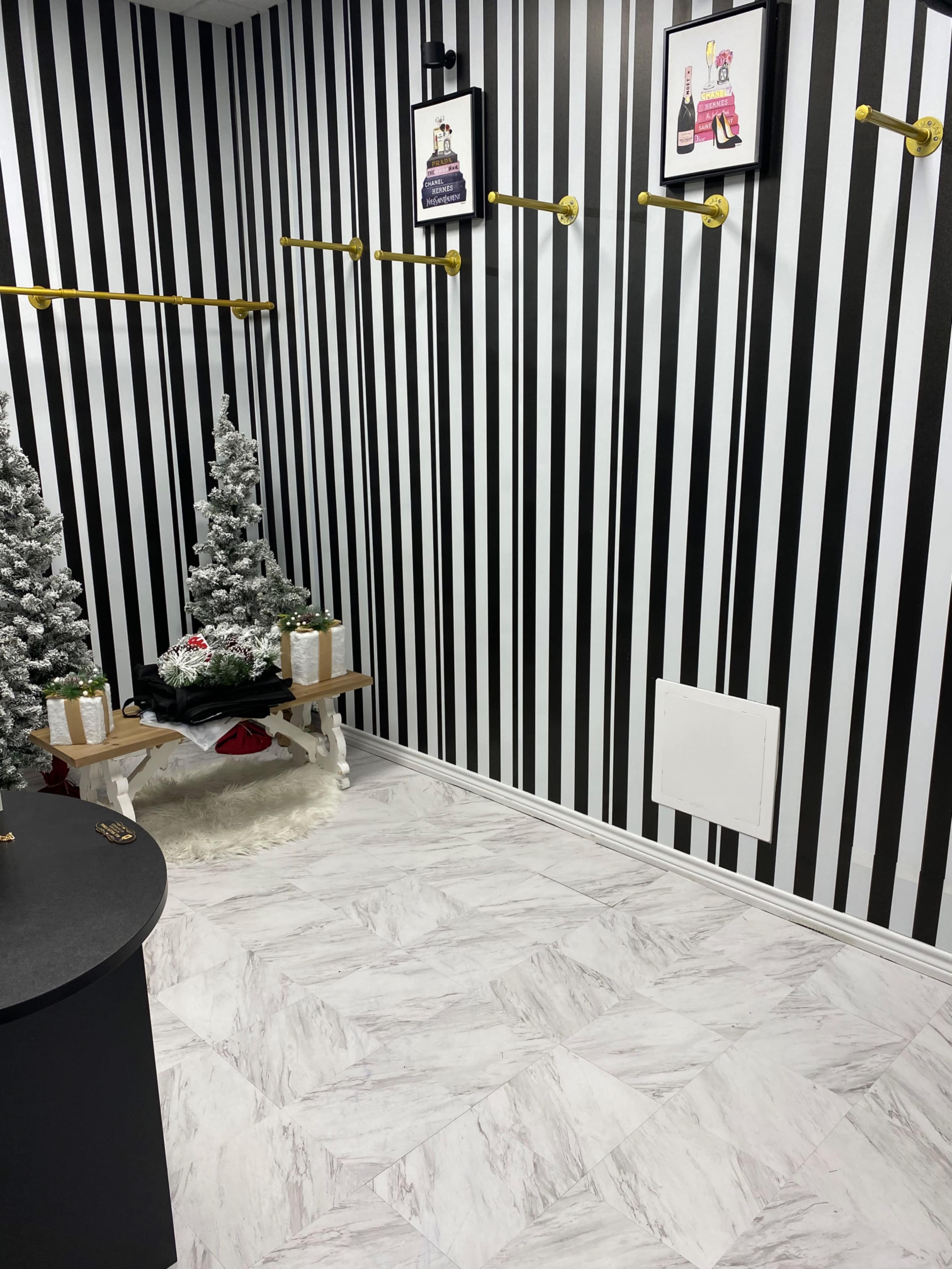 The image depicts a room with black and white striped walls, gold hangers along the walls, two small artificial Christmas trees, and a light-colored floor.