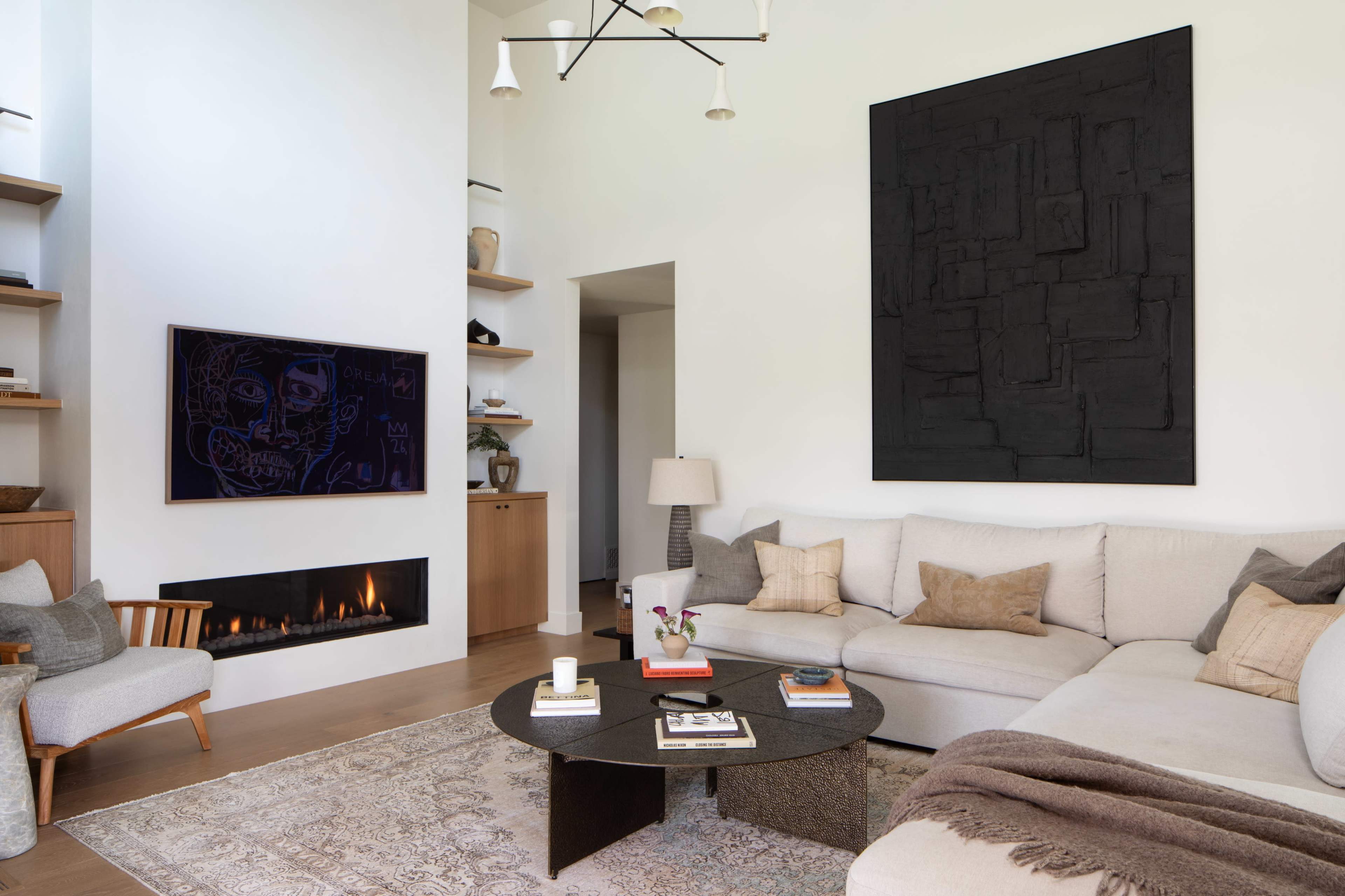 The image shows a modern living room featuring a large sectional sofa, a round coffee table, a wall-mounted television, and a fireplace, with minimalist decor and a large black artwork on the wall.