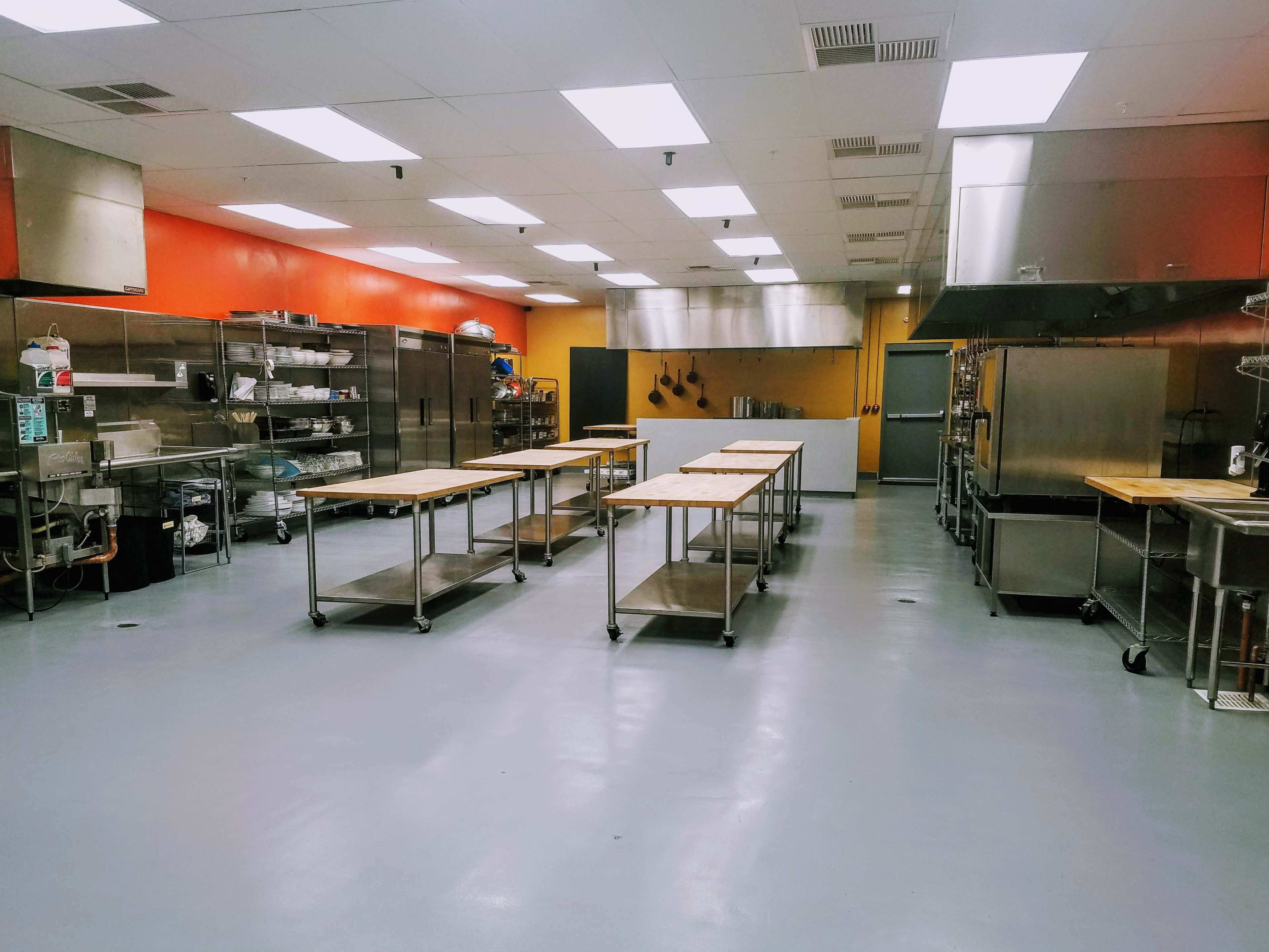 A spacious commercial kitchen with stainless steel appliances, several wooden preparation tables on wheels, and orange walls.