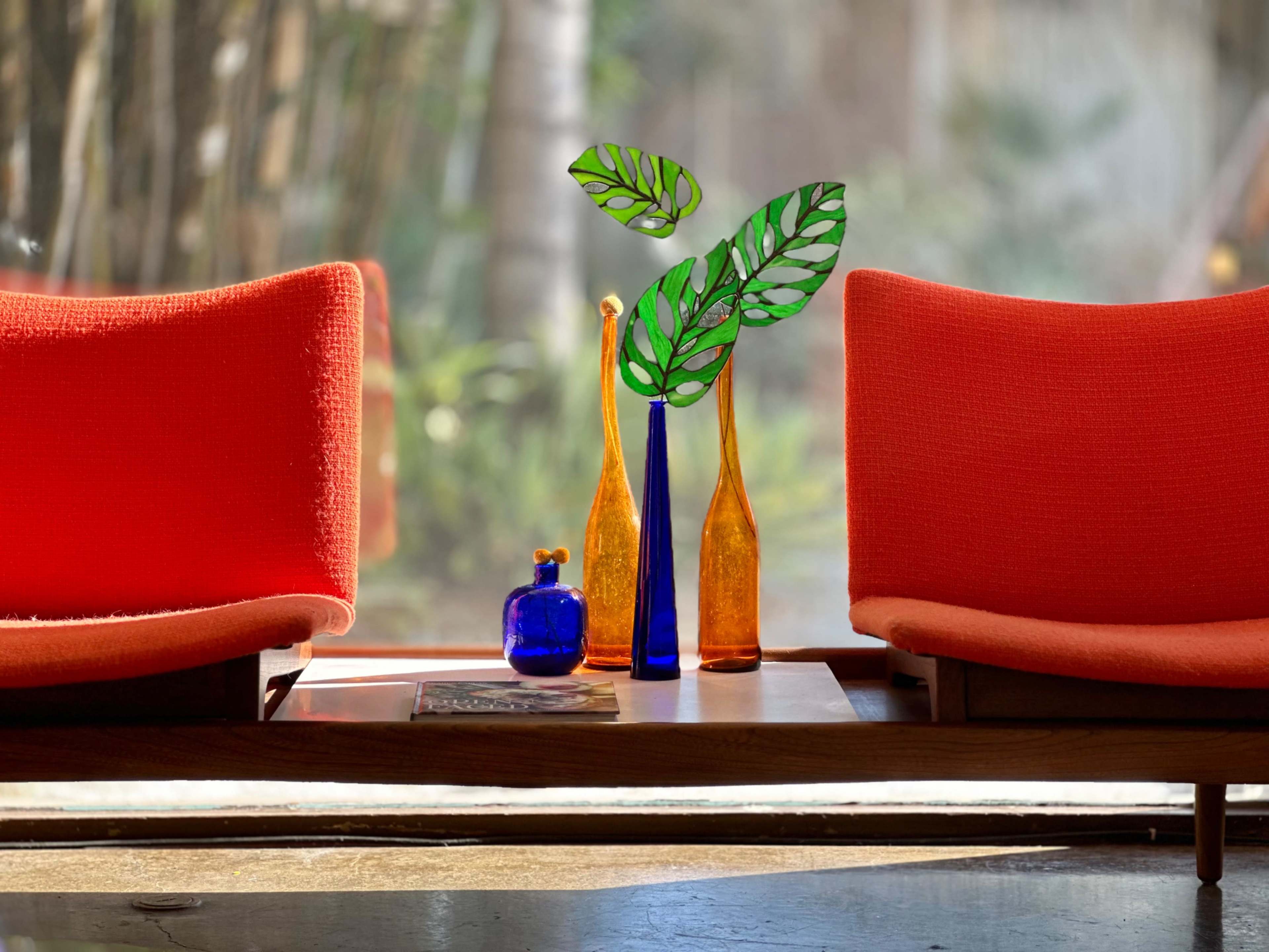 Two orange chairs frame a table displaying a colorful arrangement of glass vases and a green leaf decoration, with a blurred garden scene visible in the background.