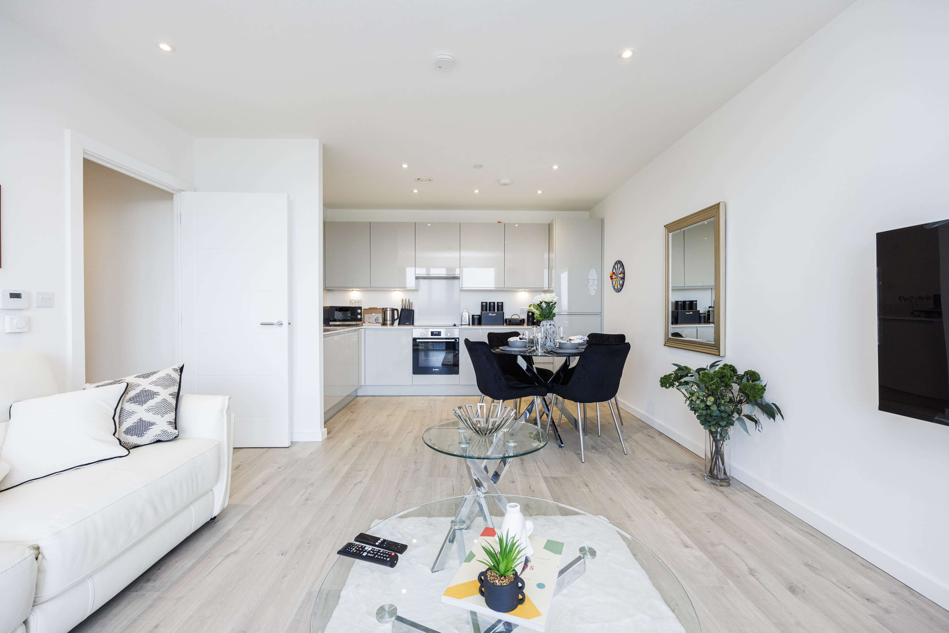 A modern living space with a white sofa, a glass coffee table, a dining area with black chairs, and a minimalist kitchen in the background.