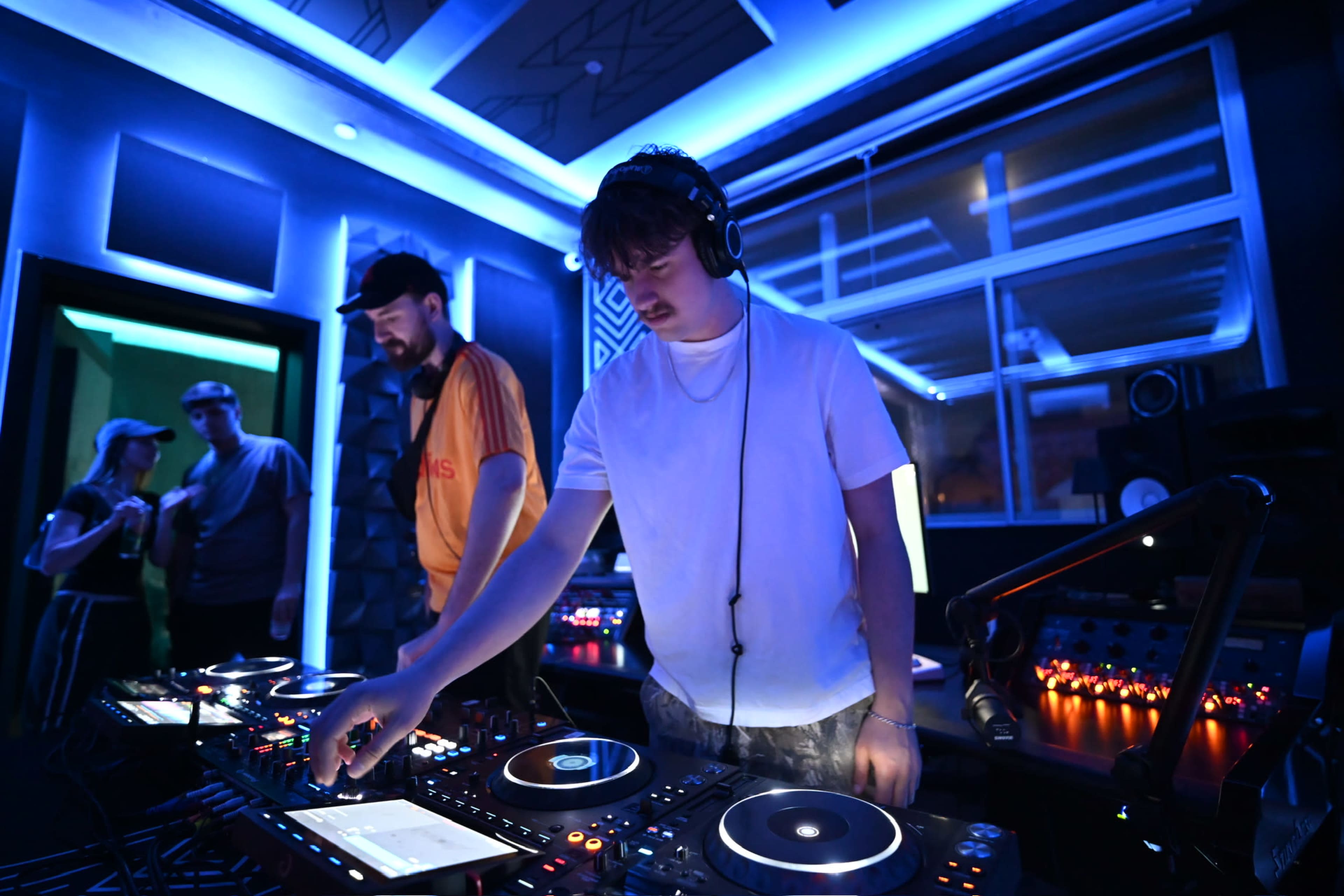 A DJ is mixing music on turntables in a dimly lit room with blue lighting, while two people observe nearby.