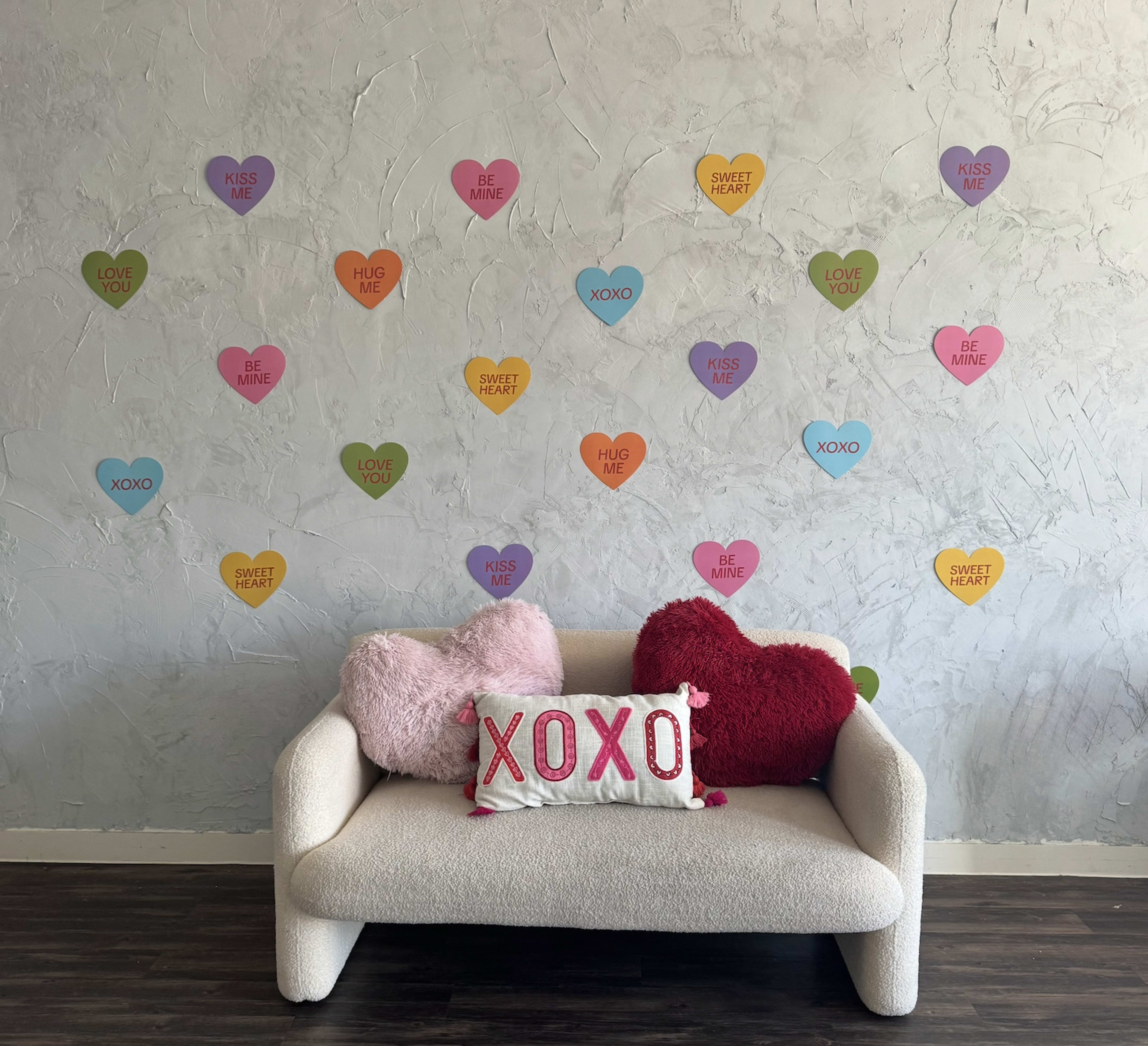 A cozy couch with decorative pillows is set against a textured wall, adorned with colorful heart-shaped decorations featuring various affectionate phrases.