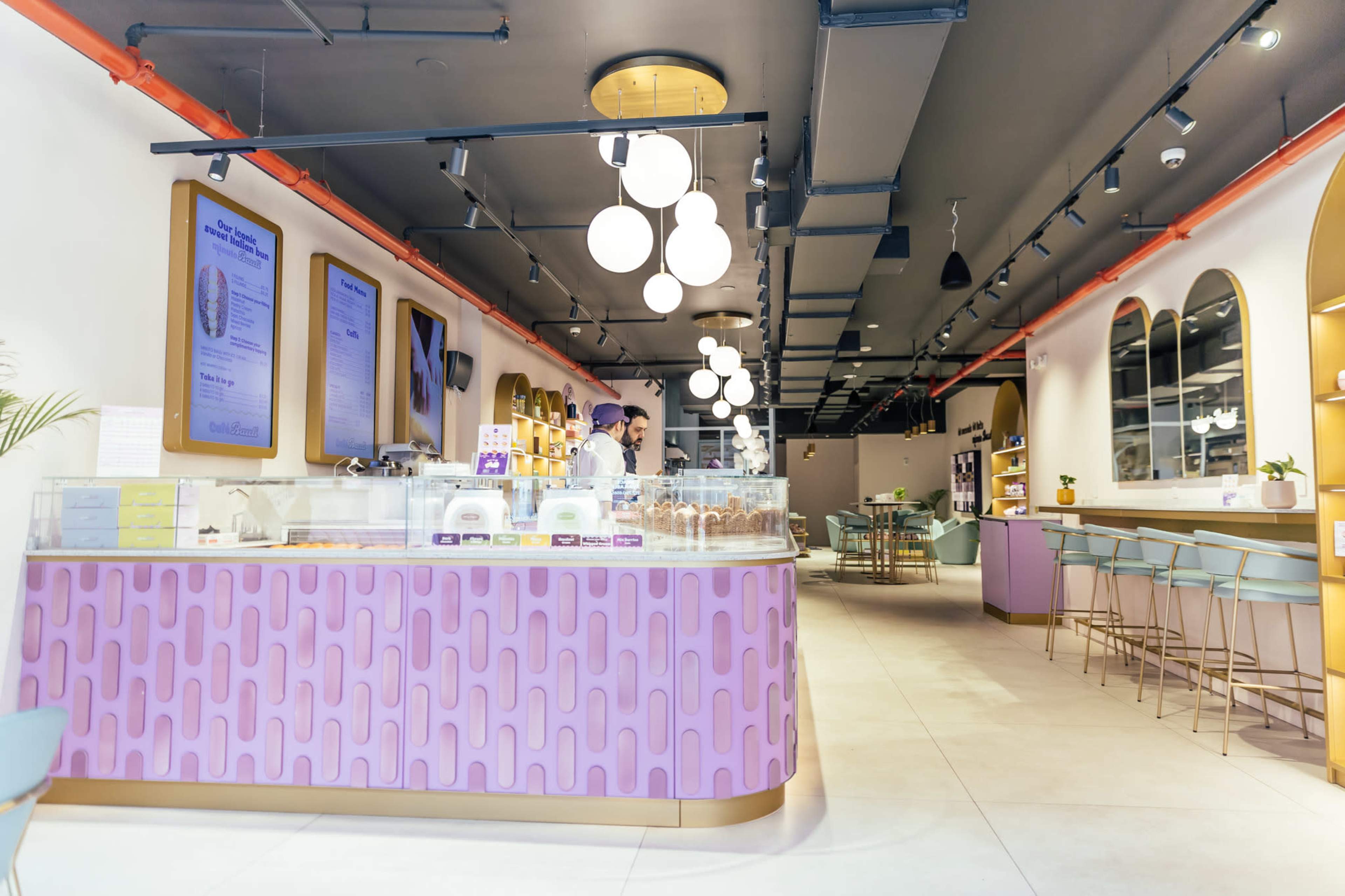 The image shows a modern café interior with a pink service counter, seating areas, and decorative lighting.