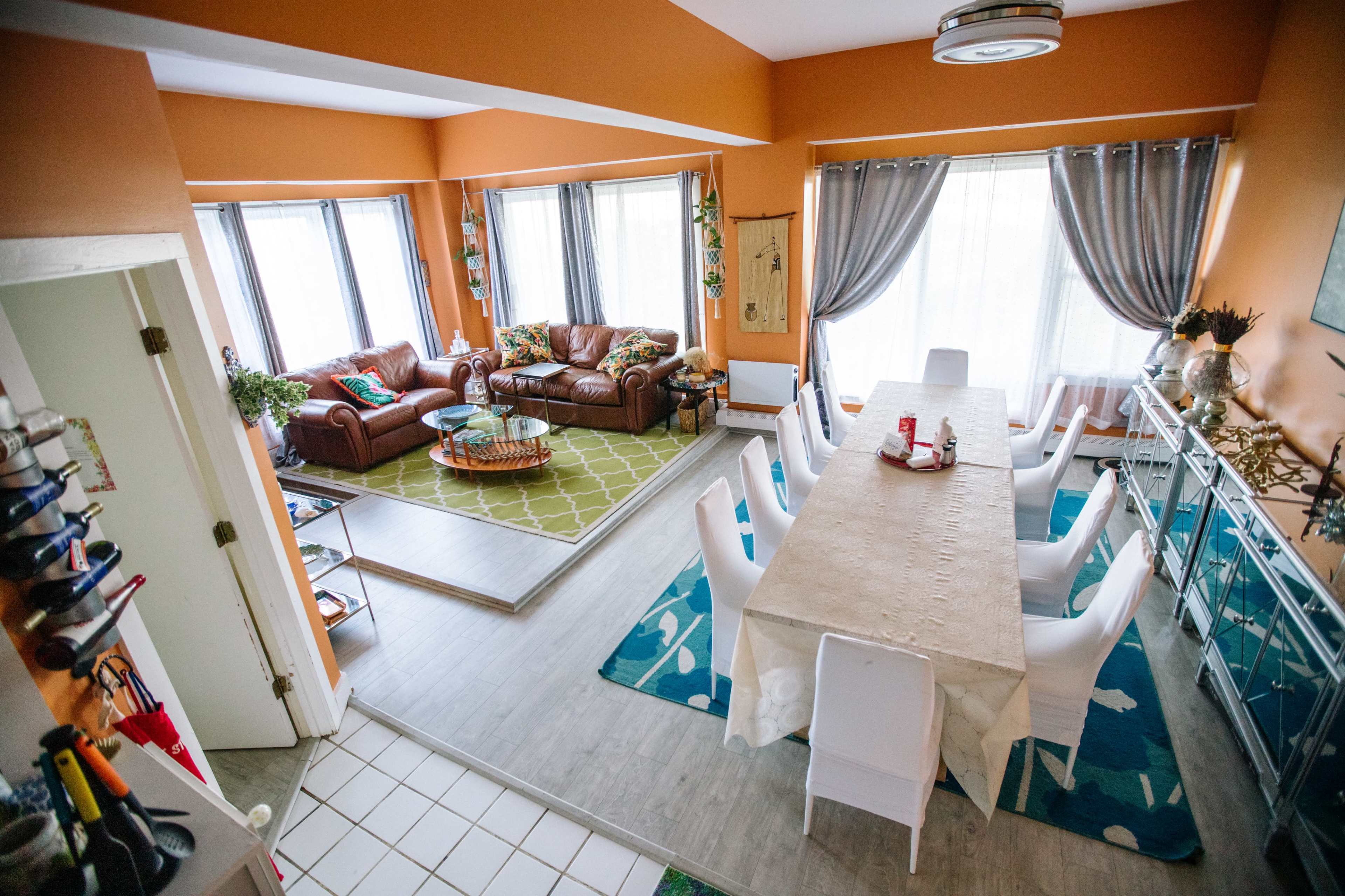 The image shows a spacious, open-concept living and dining area with a large dining table, comfortable seating, and large windows allowing natural light.