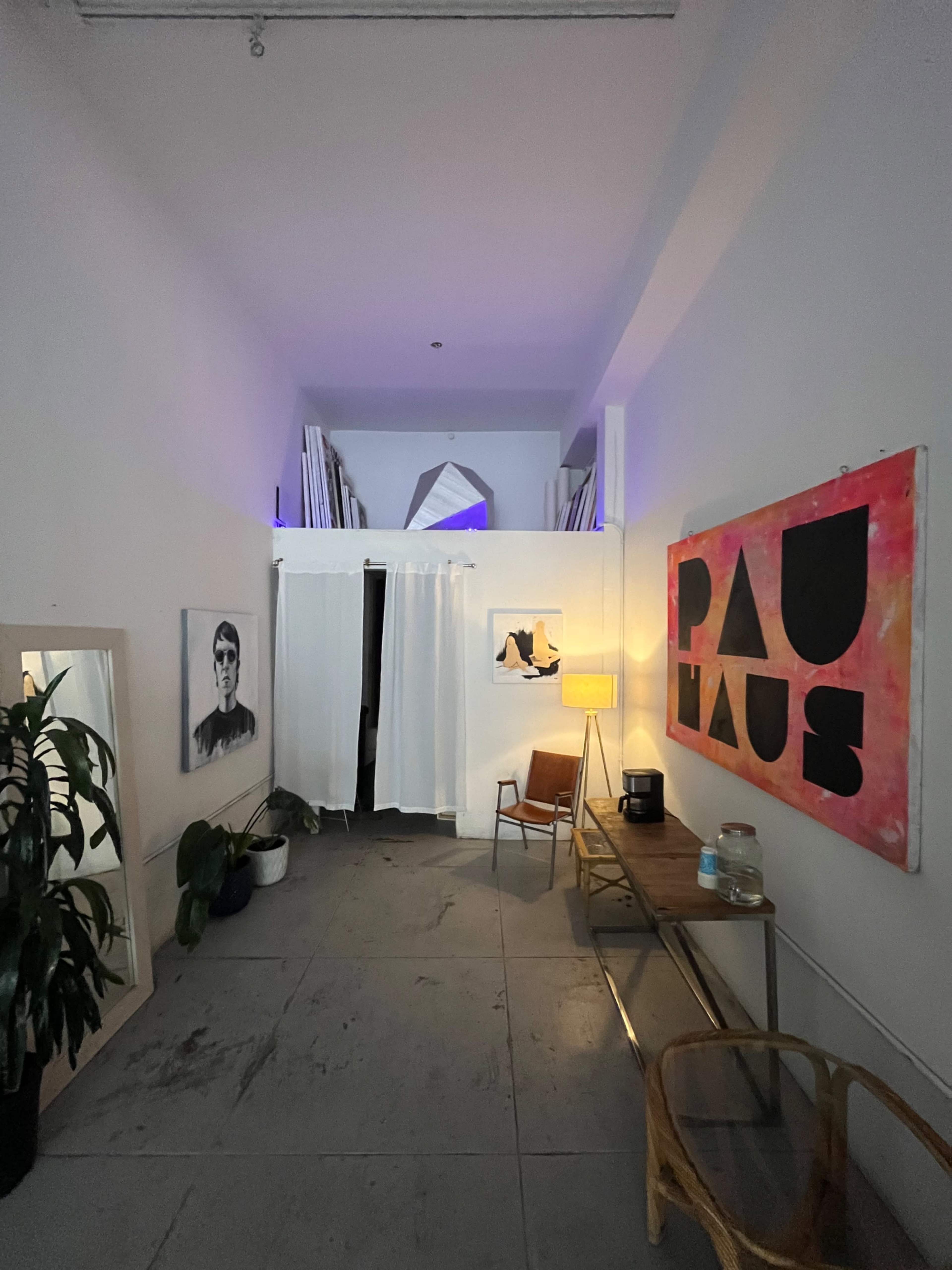 A minimalist interior space with white curtains, artworks on the walls, a small table, a chair, and a plant.