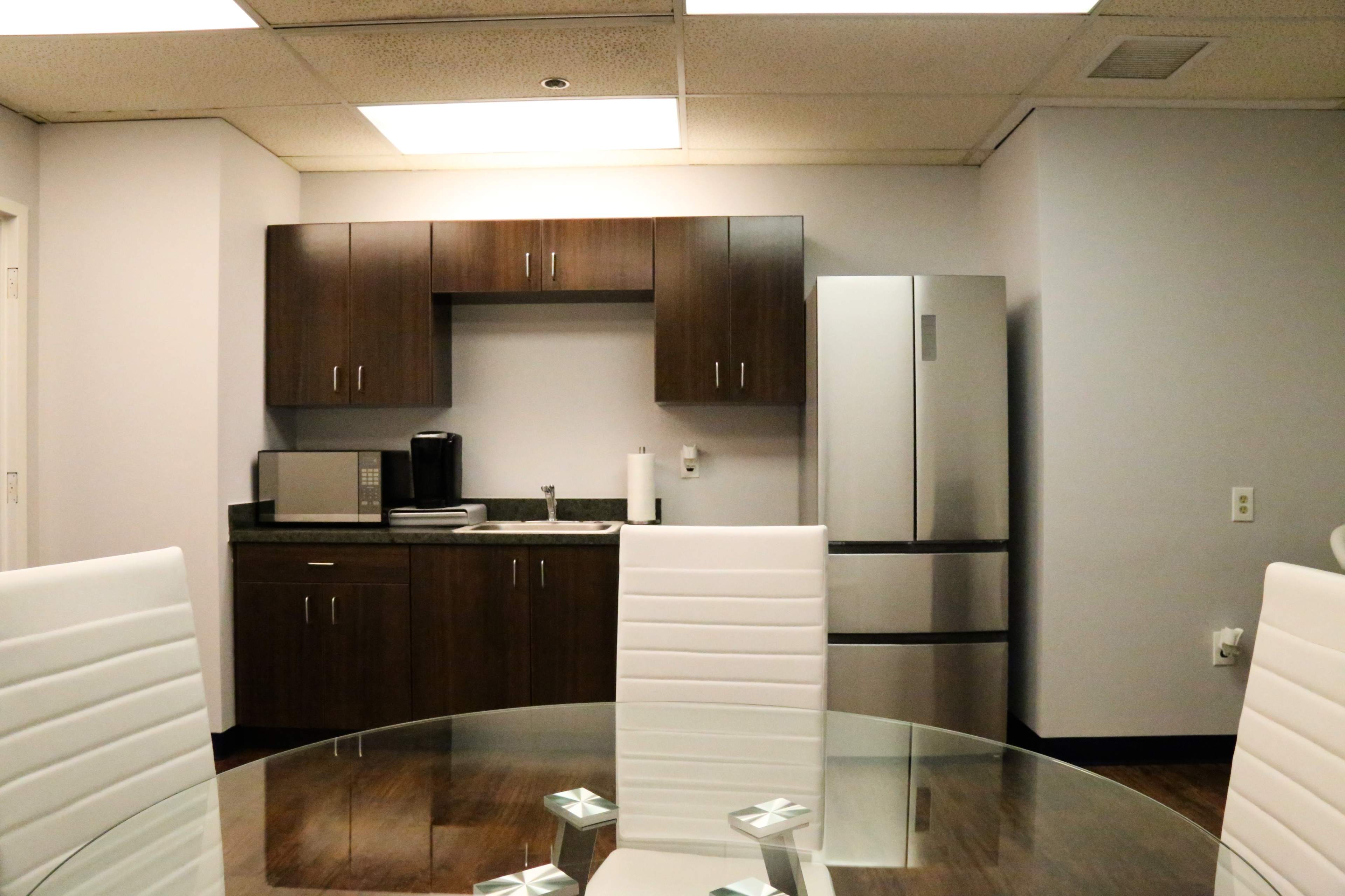 A modern kitchenette with a microwave, coffee maker, and a refrigerator, set against a backdrop of light-colored walls and a glass dining table with white chairs.