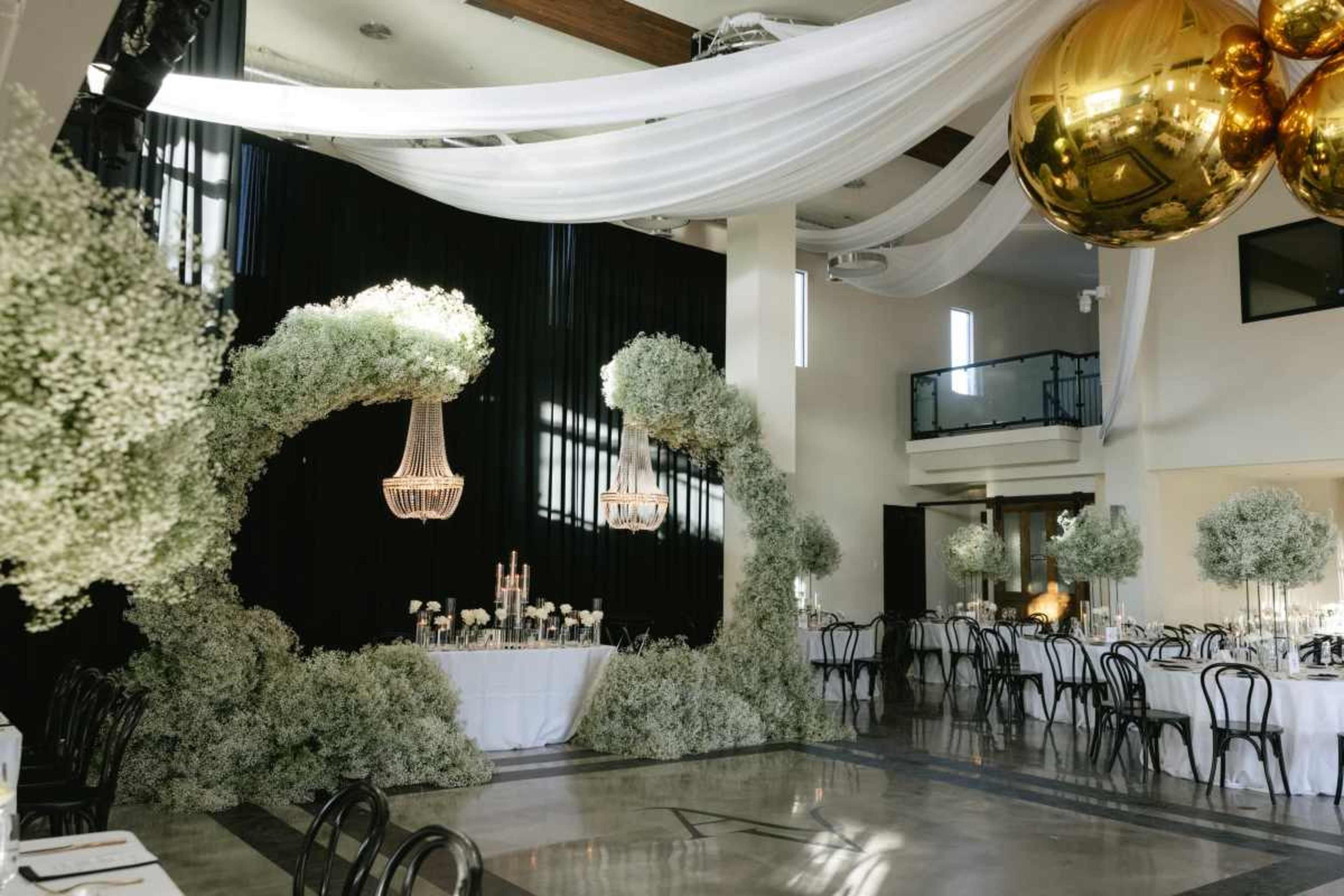 The scene features a decorated event space with floral arrangements, chandeliers, and a simple table setup surrounded by black and gold decor elements.