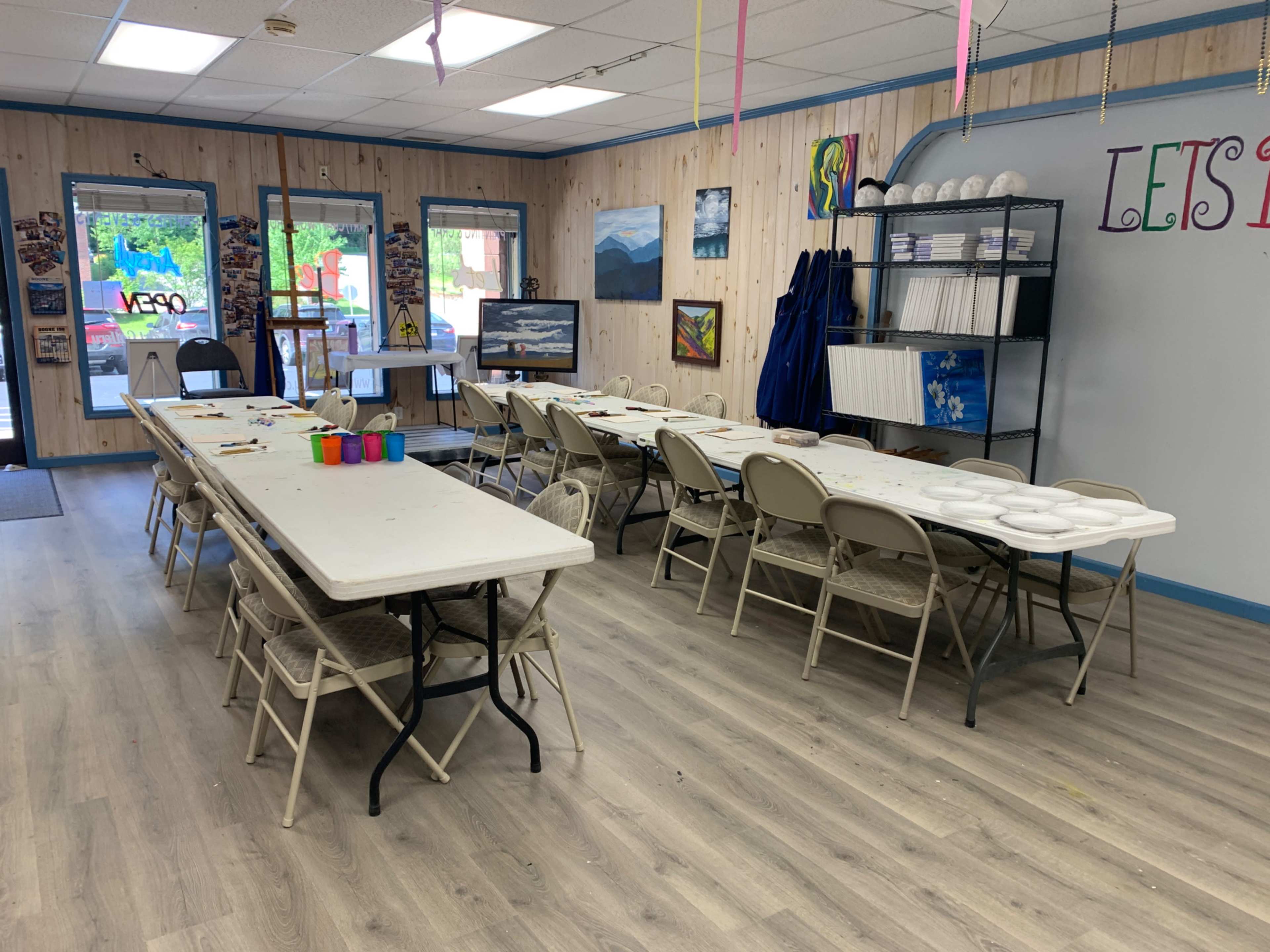 The image shows a bright, organized art studio with several tables and chairs set up for painting activities, along with art supplies and finished works displayed on the walls.