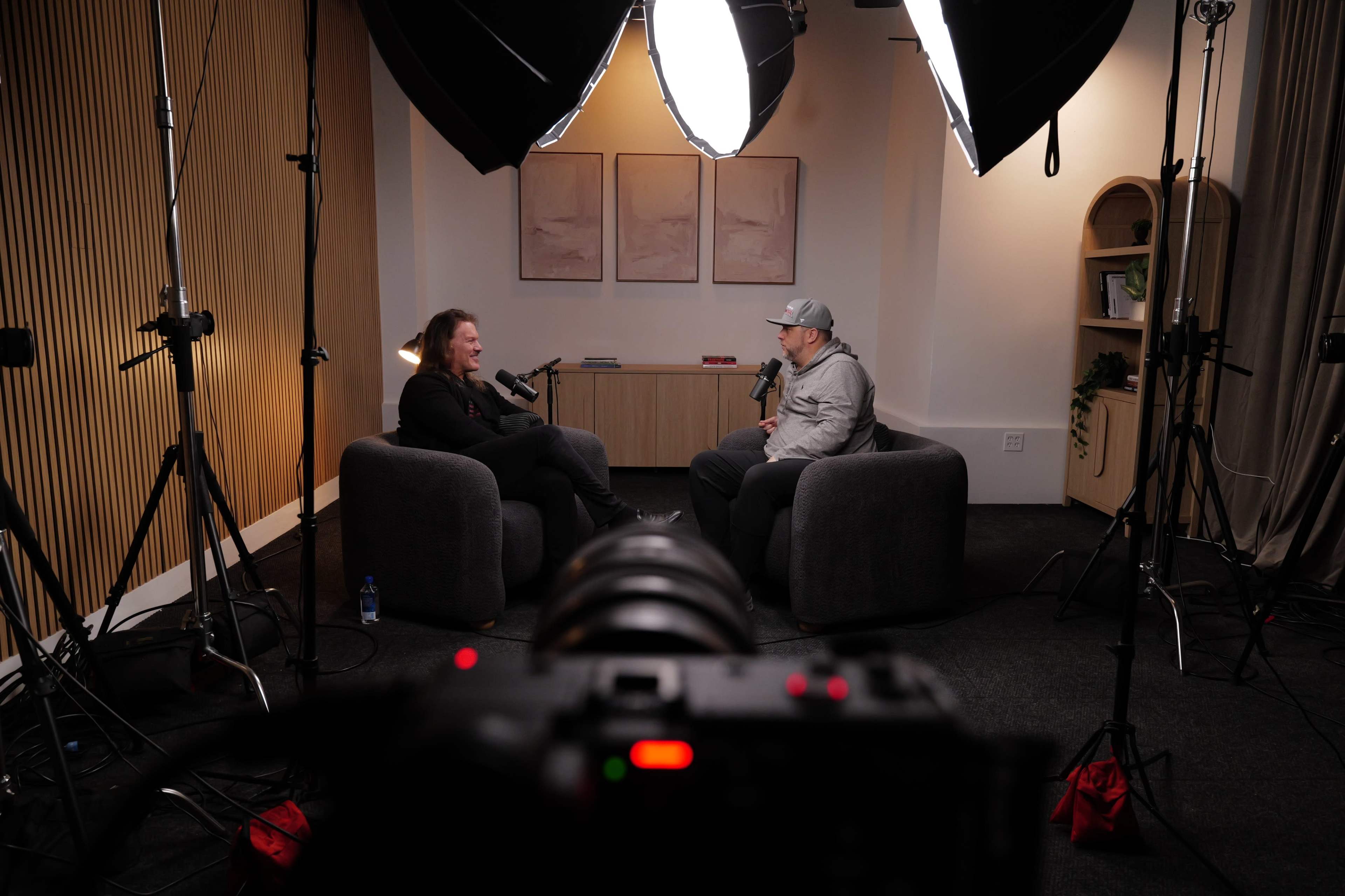 Two men are seated in armchairs during a podcast recording in a well-lit studio, surrounded by cameras and lighting equipment.
