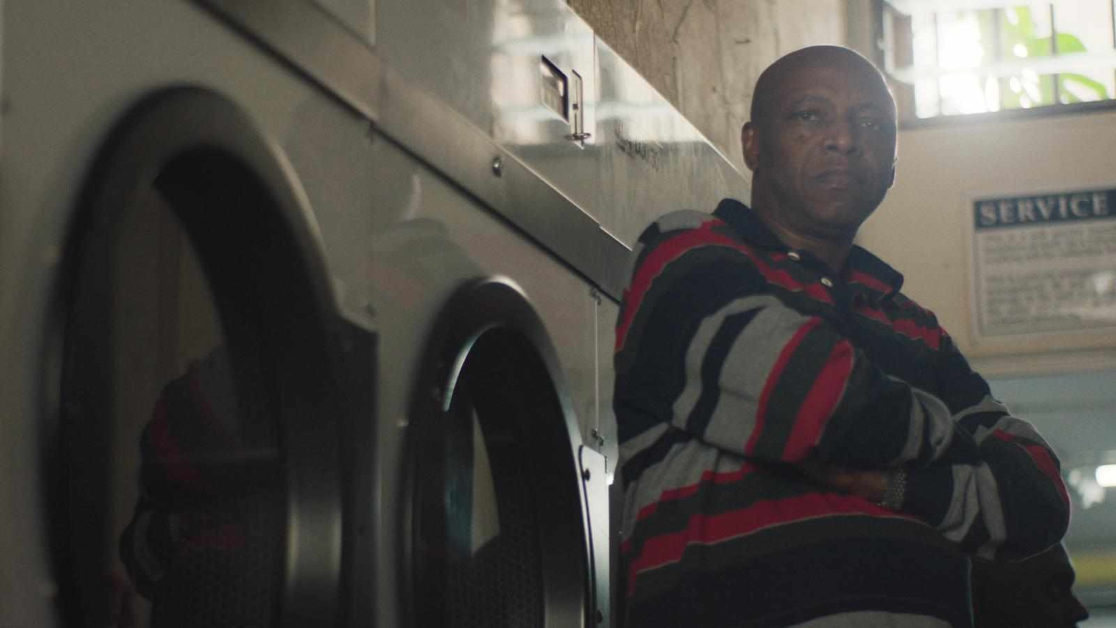 A man in a striped shirt stands with his arms crossed next to a row of washing machines in a laundry facility.