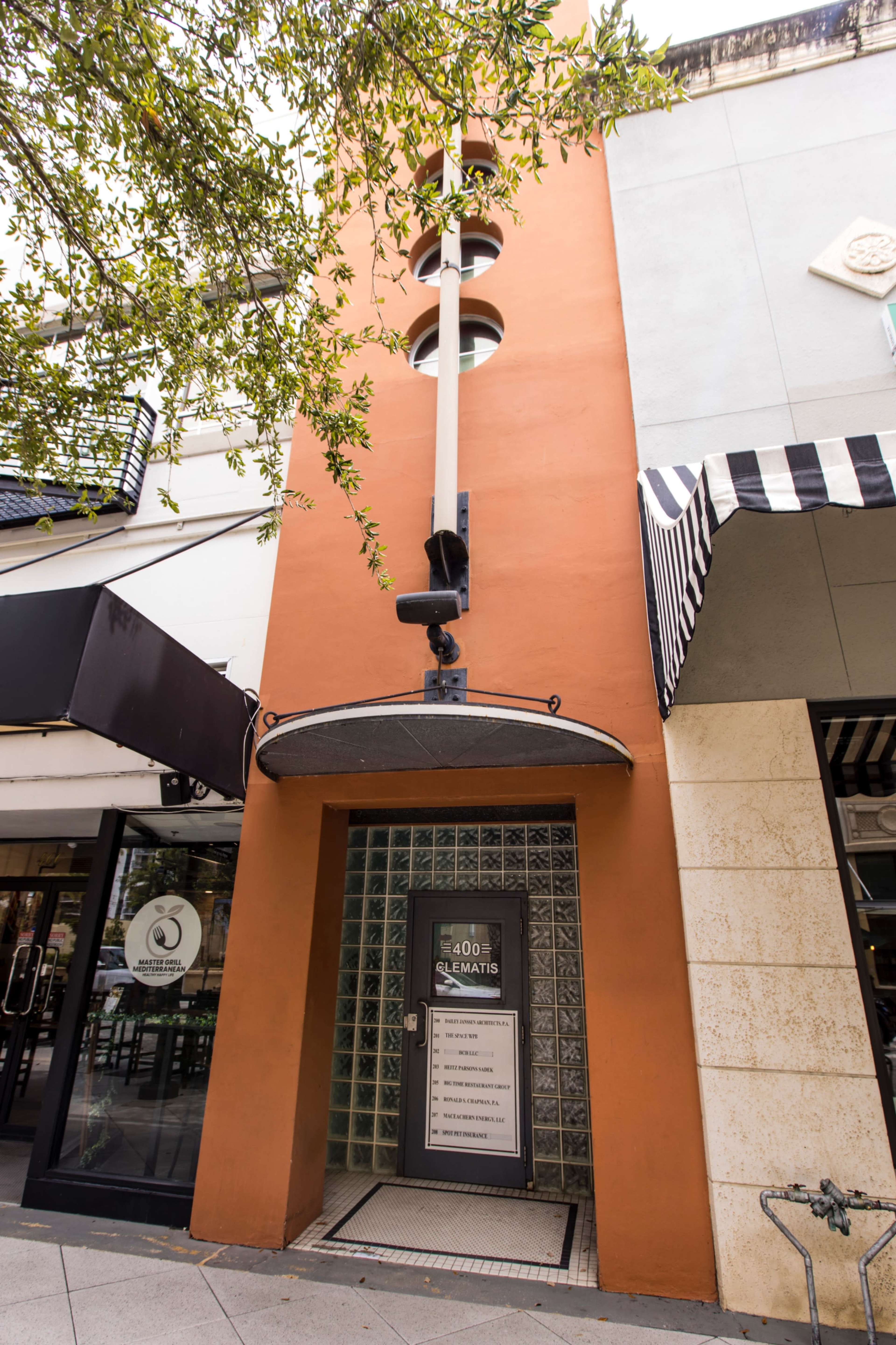The image shows an orange building with a glass front and a large awning extending over a door that features a sign with various business names listed on it.